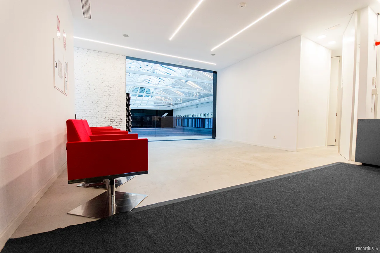 A minimalist reception area features a row of bright red armchairs on a light concrete floor next to an exposed white brick wall.