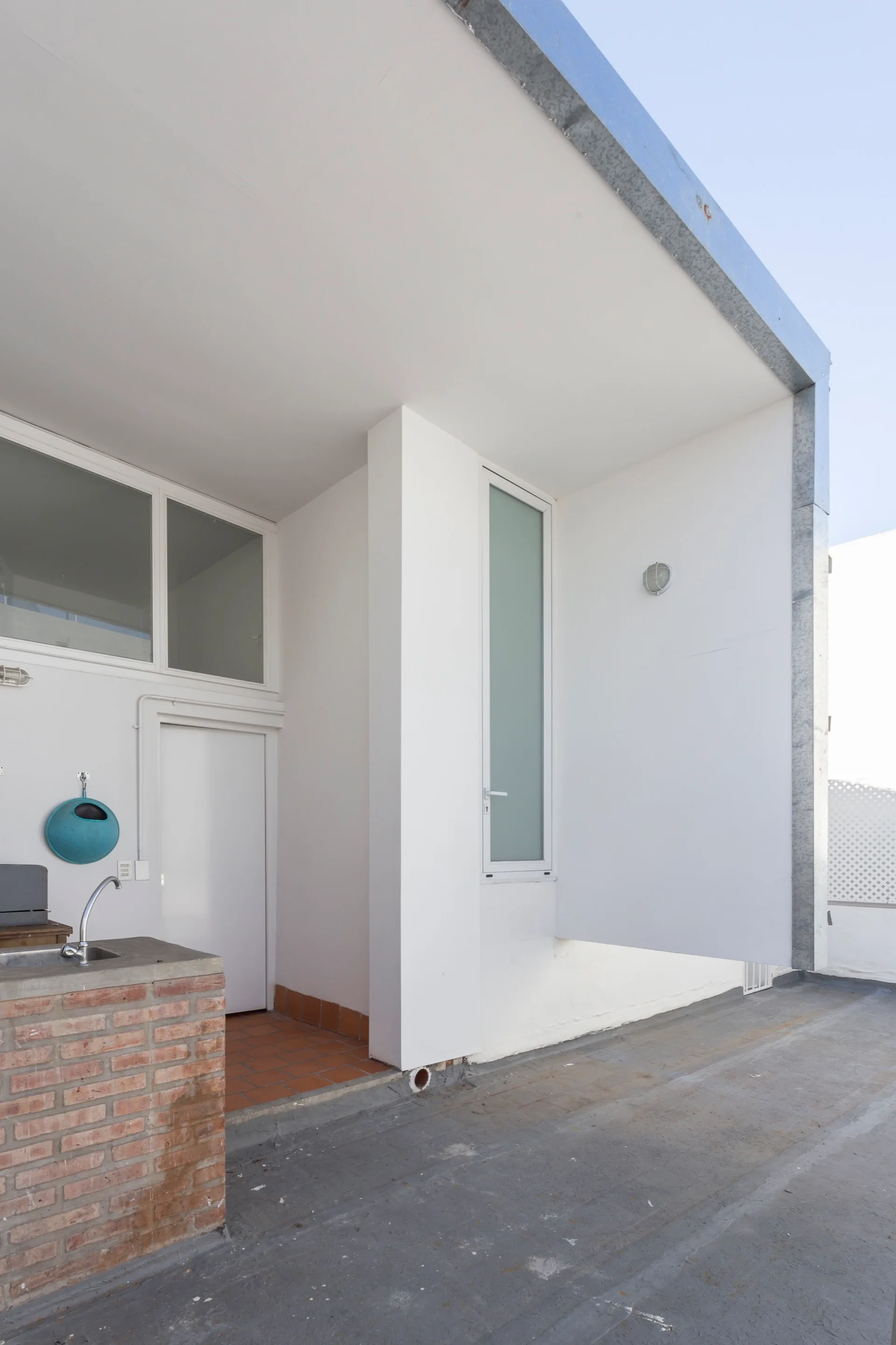 A minimalist outdoor terrace features smooth white stucco walls, a gray concrete floor, and a compact utility sink with a red brick base.