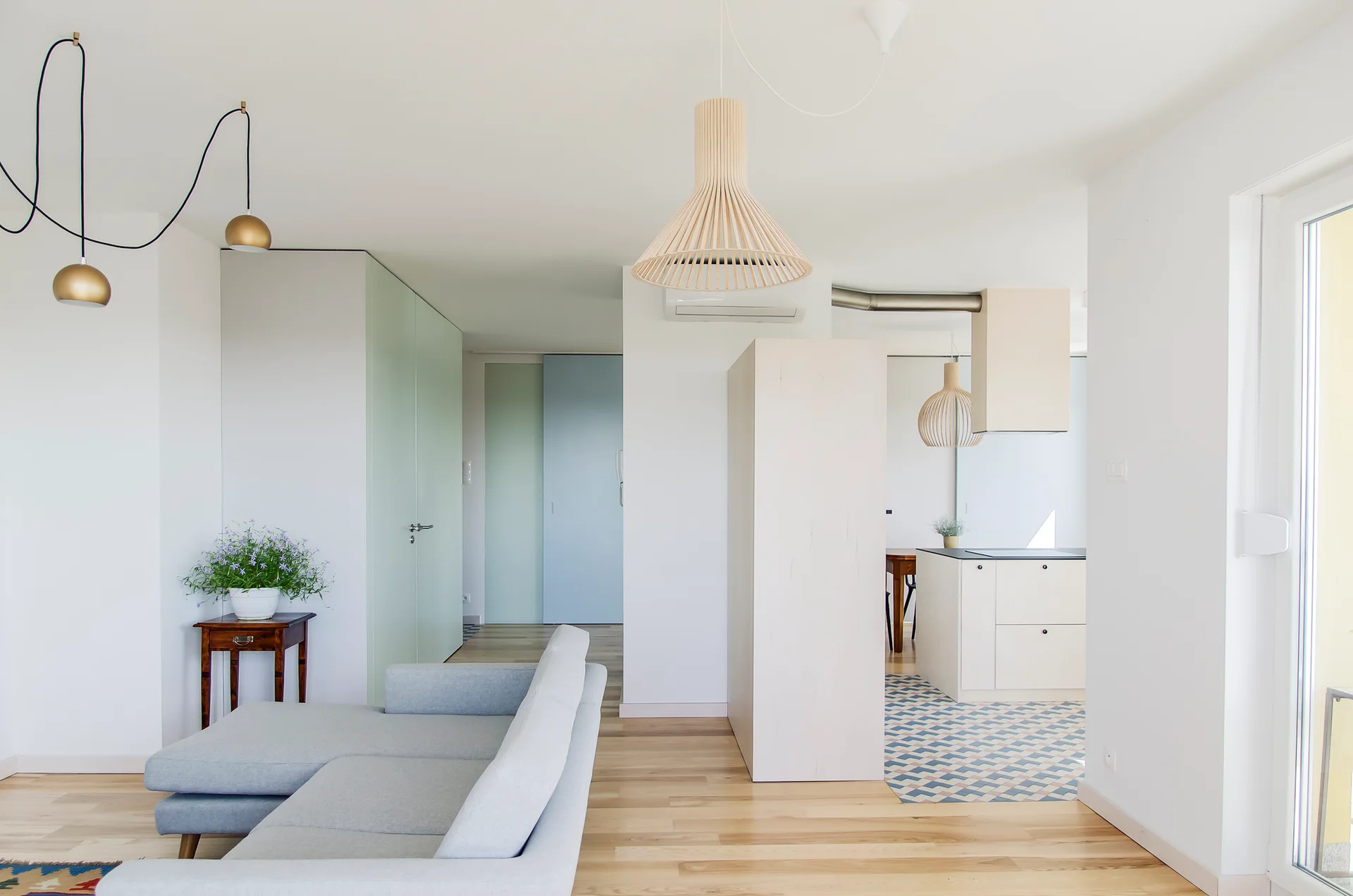A light-filled open-plan living room features a grey sectional sofa, pale green and blue sliding doors, and a kitchen with geometric floor tiles.