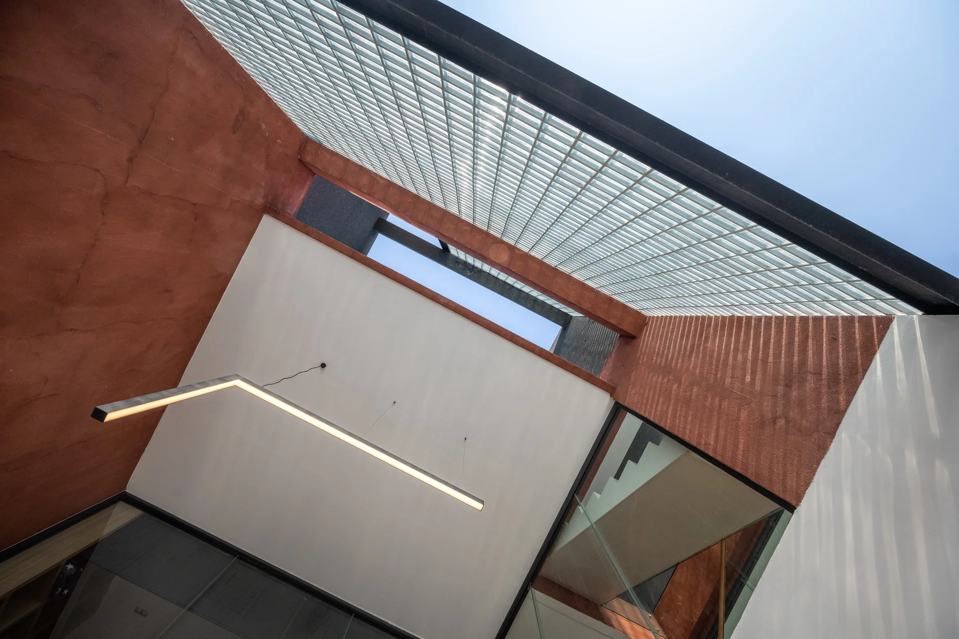 An upward view of a modern stairwell featuring textured terracotta plaster walls, a geometric pendant light, and a grid glass skylight.