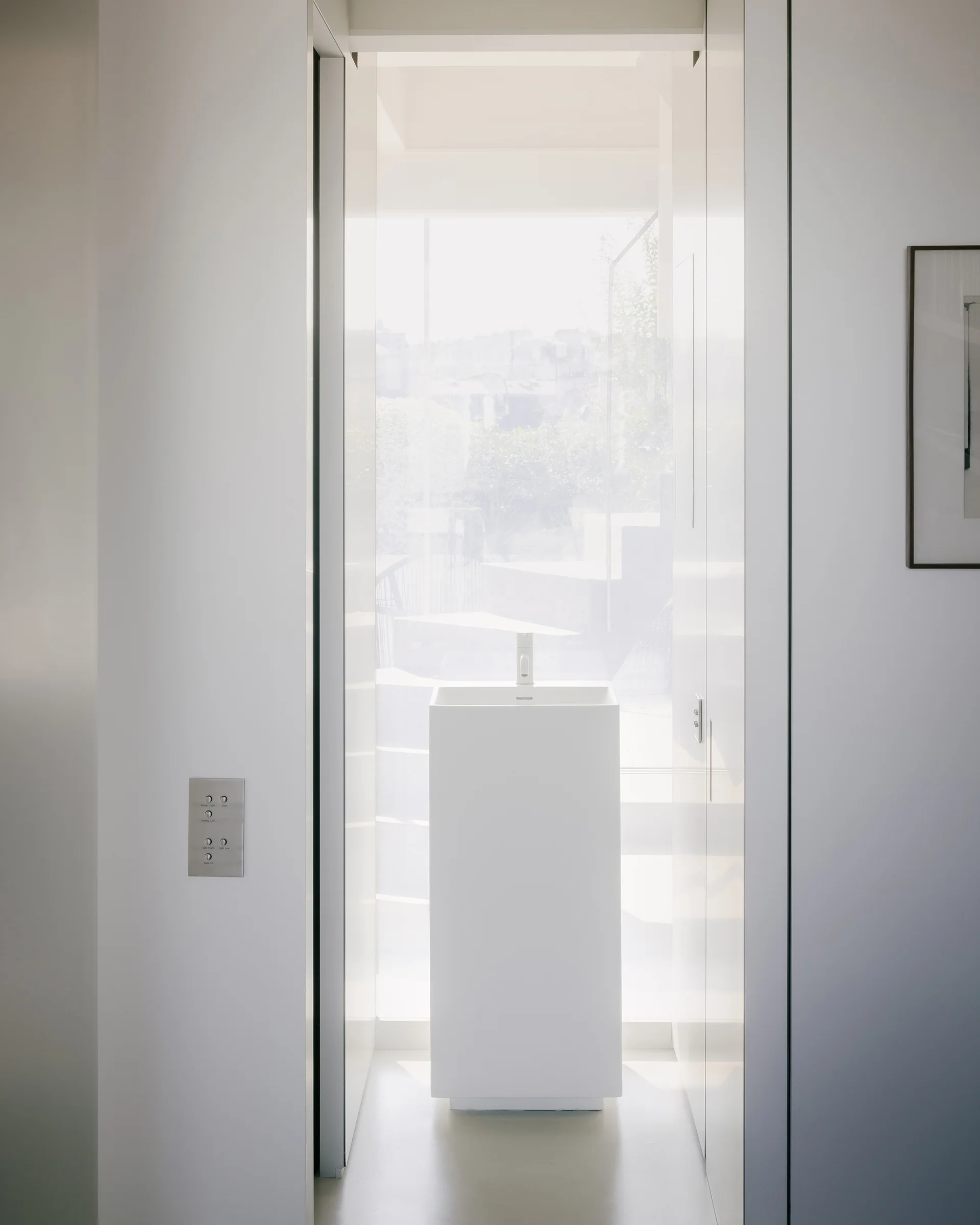 A rectangular white pedestal sink stands centered in a bright entryway niche against a large window between glossy white wall panels.