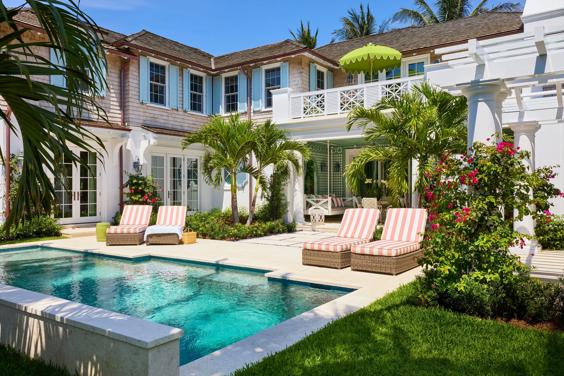A backyard swimming pool sits next to a cedar-shingled house with light blue shutters, stone paving, and striped lounge chairs.