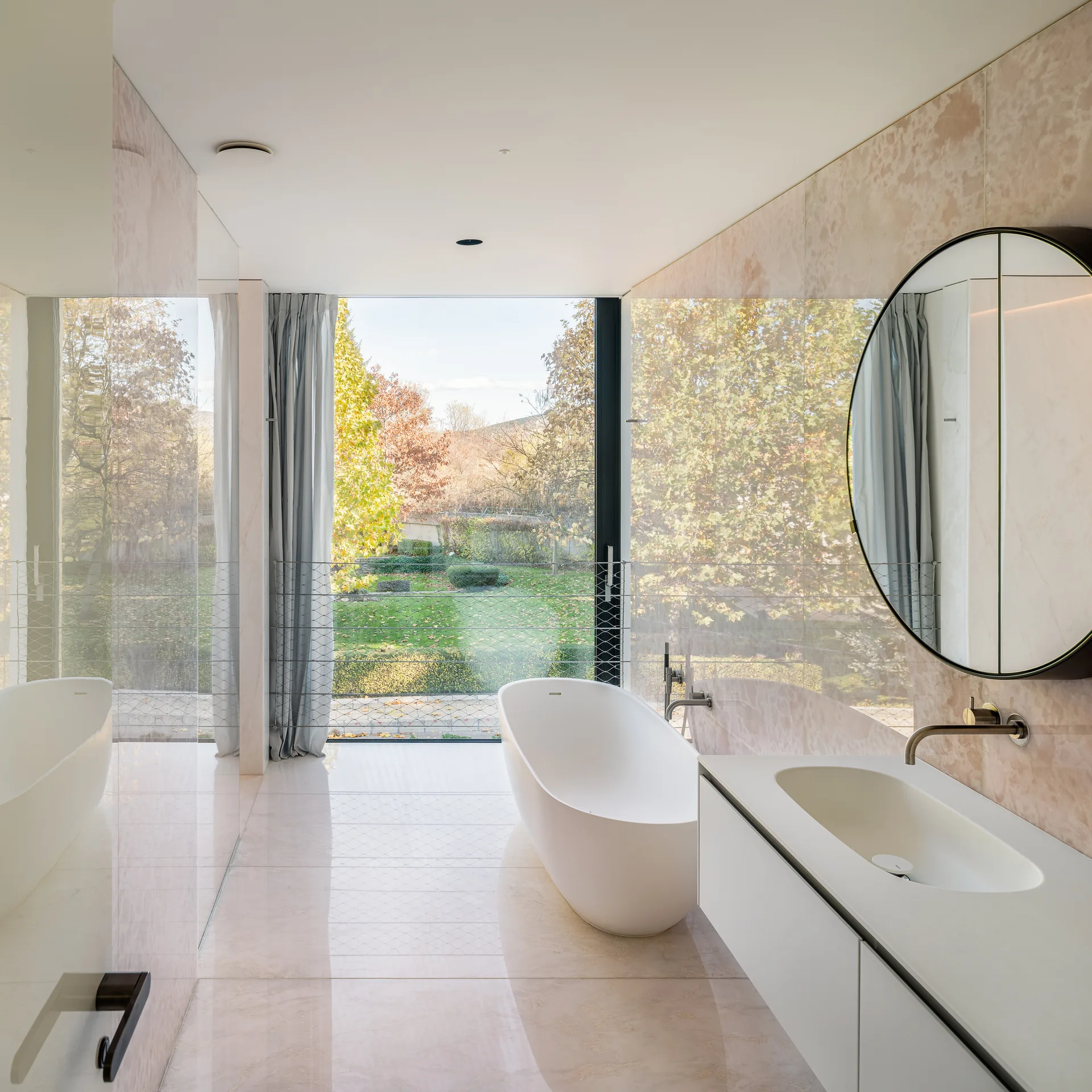 A minimalist bathroom featuring a white freestanding tub, a round mirror over a white vanity, and large beige stone wall panels facing a garden window.