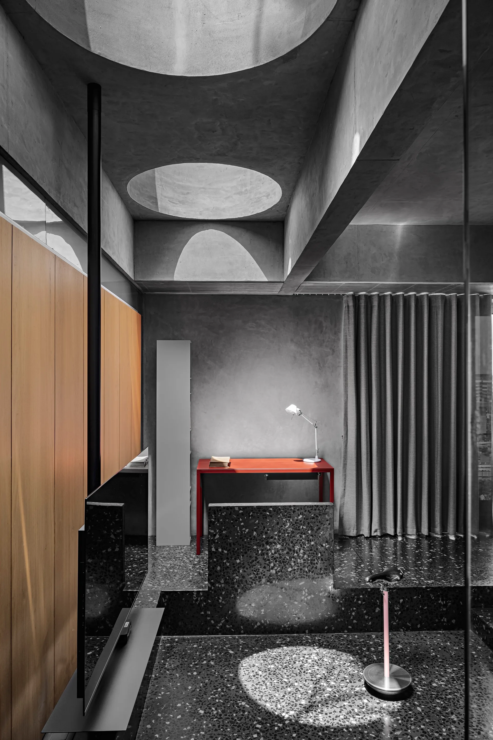 A modern minimalist study featuring a red desk on a dark speckled terrazzo floor under a raw concrete ceiling with circular skylights.