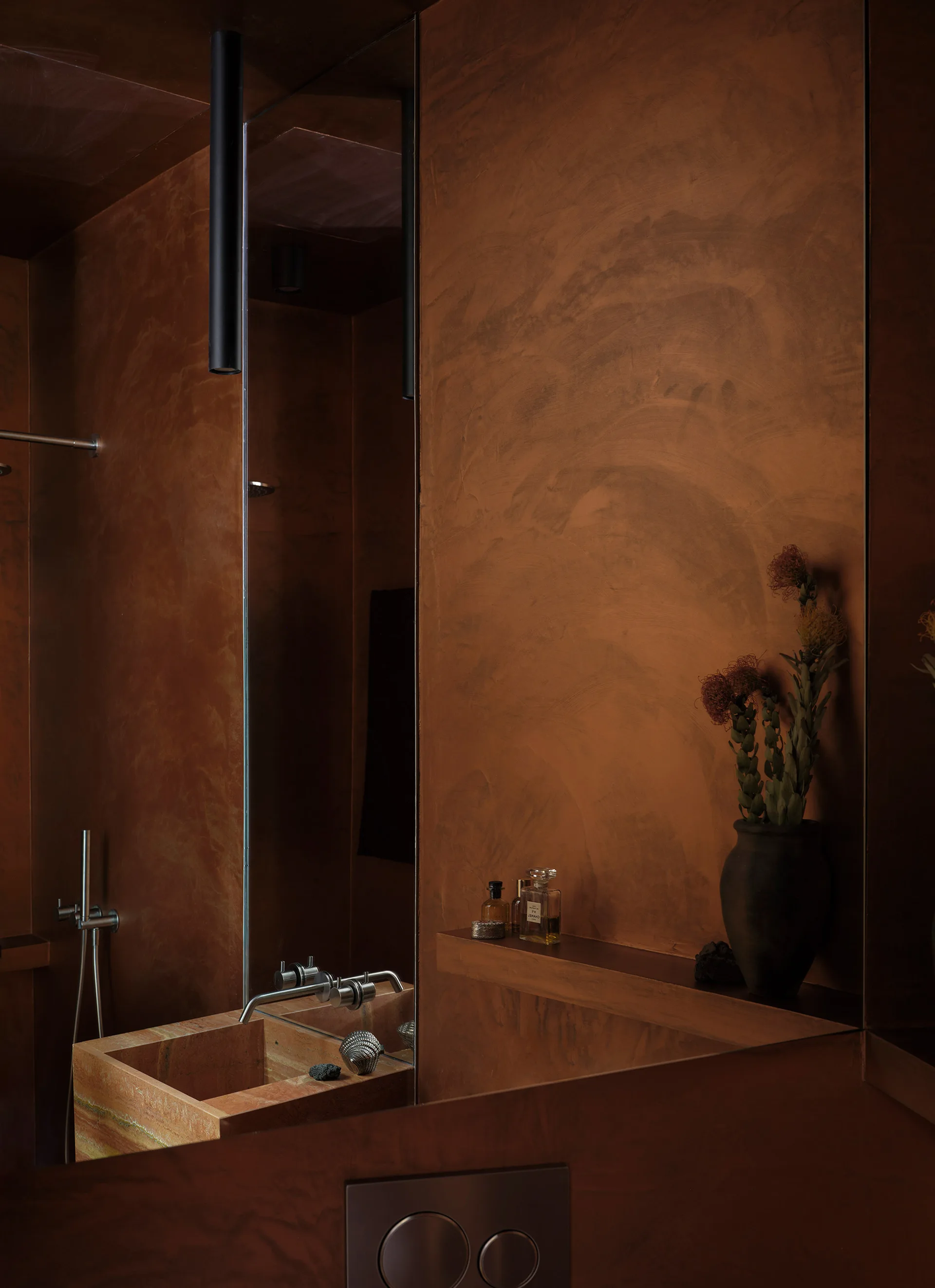 A dark brown plaster bathroom features a rectangular stone basin sink, a wall-mounted faucet on a vertical mirror, and a small built-in shelf.