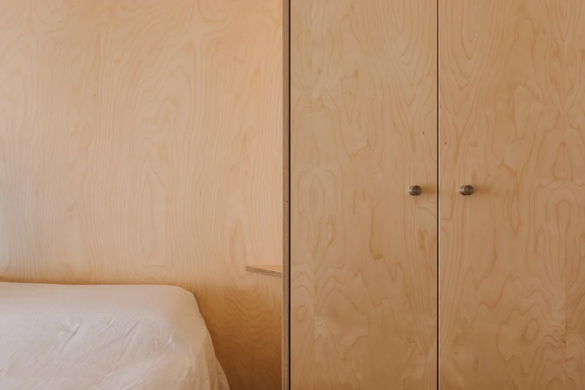 A minimalist bedroom detail features natural light plywood wall paneling, a matching wardrobe with round metal knobs, and a bed with white sheets.