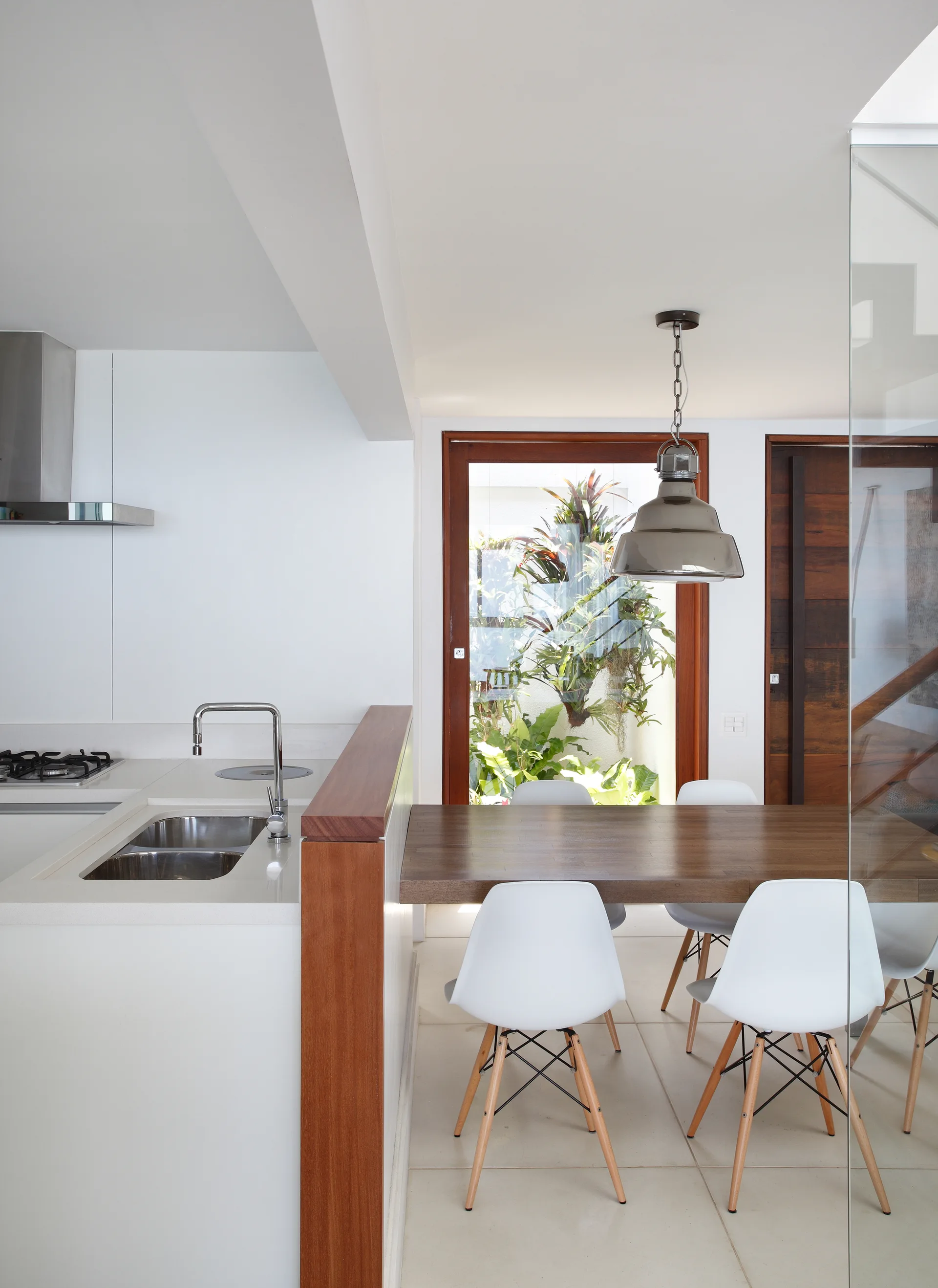 A modern white kitchen features a dark wood dining table, white molded chairs, a glass partition, and a view of a garden through a back window.