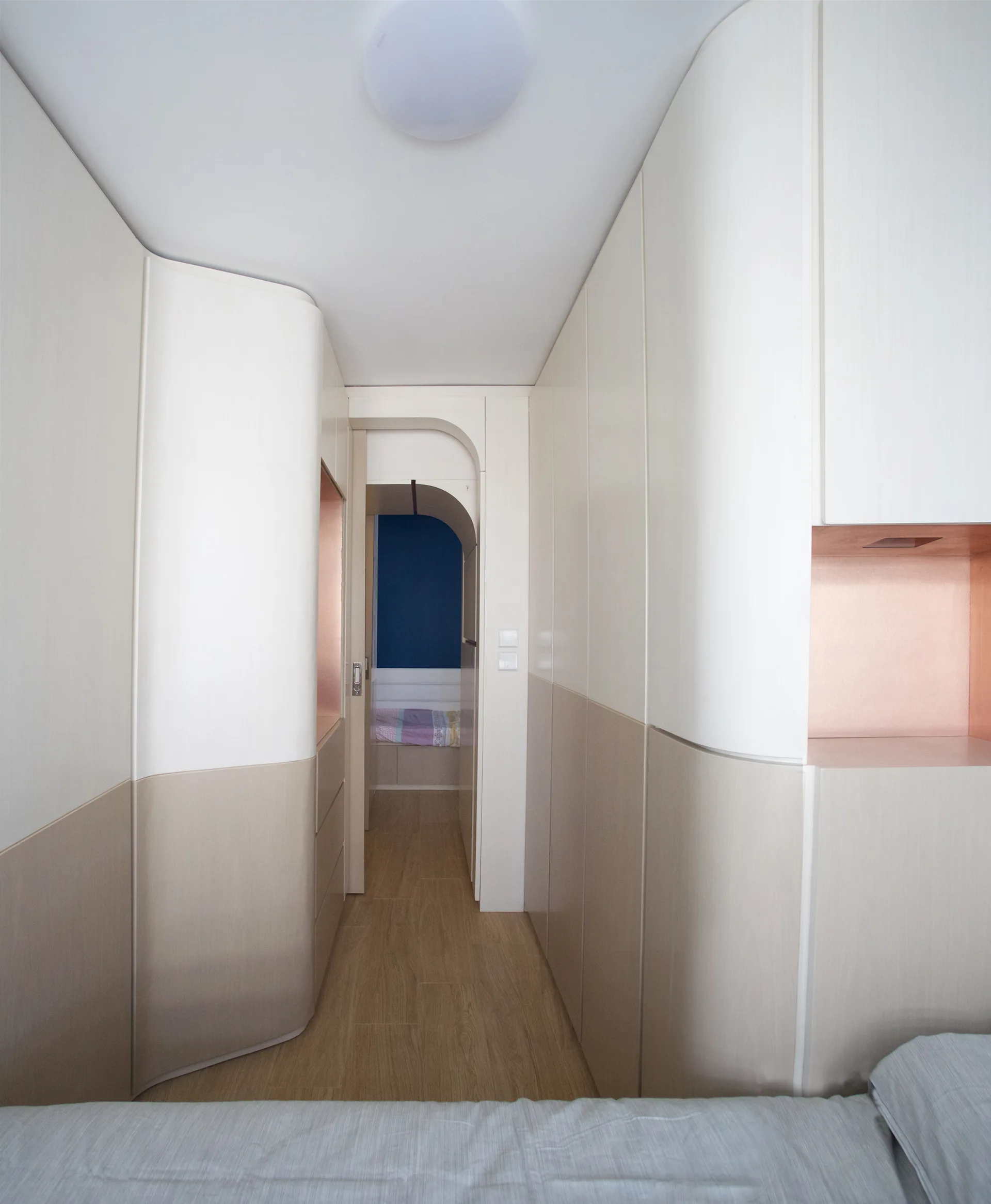 A narrow hallway featuring curved floor-to-ceiling built-in cabinetry in cream and beige with recessed copper niches leading to an arched doorway.