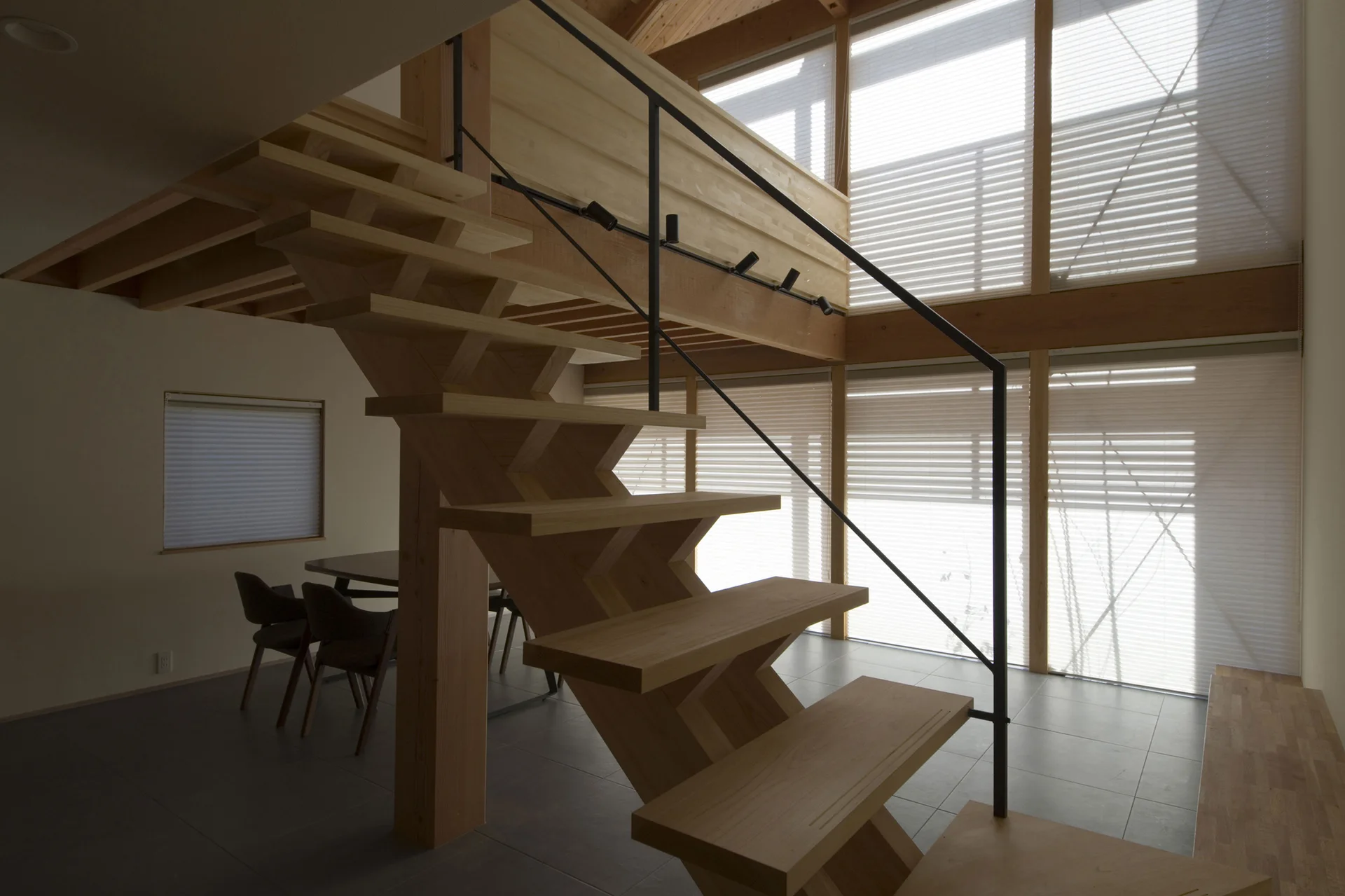 A wooden zig-zag staircase with a thin black metal railing stands before a double-height wall of windows in a modern home.