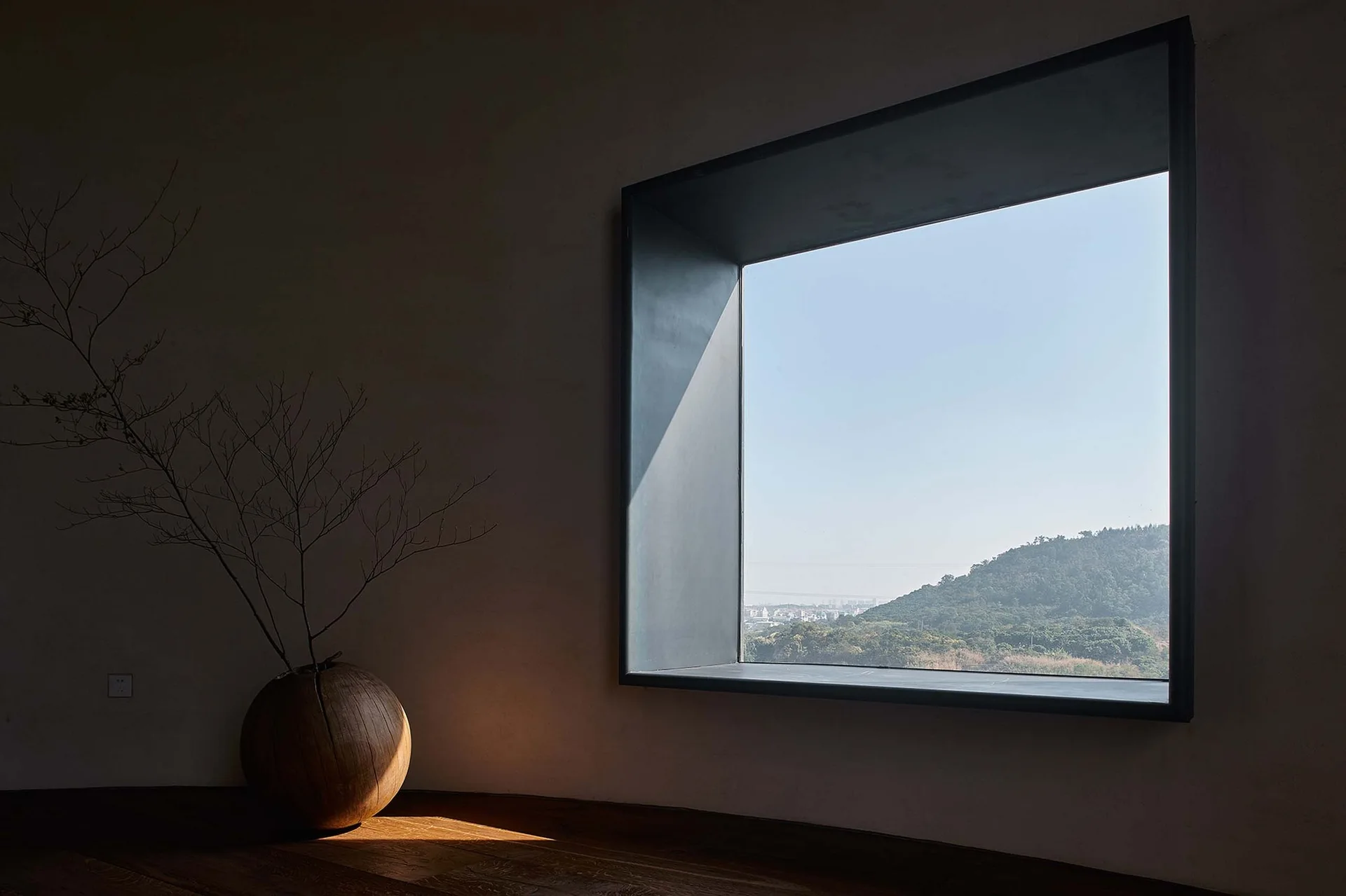 A minimalist dark room features a deep-set square window framing a green hill, with a round wooden vase holding a dry branch on the floor.