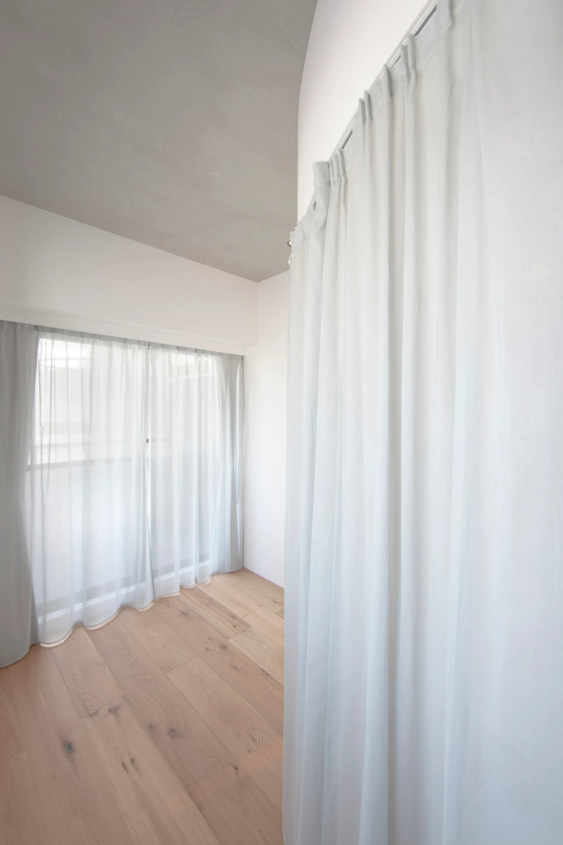 A minimalist room corner with light wood floors, smooth white walls, and floor-to-ceiling sheer white curtains hung from a track on the ceiling.