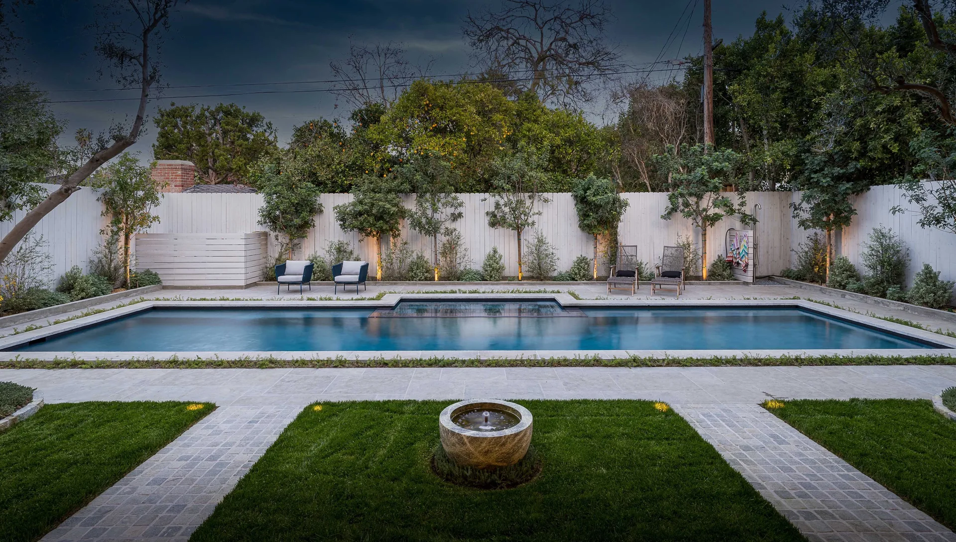 A rectangular swimming pool is surrounded by stone pavers, green lawn sections, a stone fountain bowl, and a tall white wooden fence.