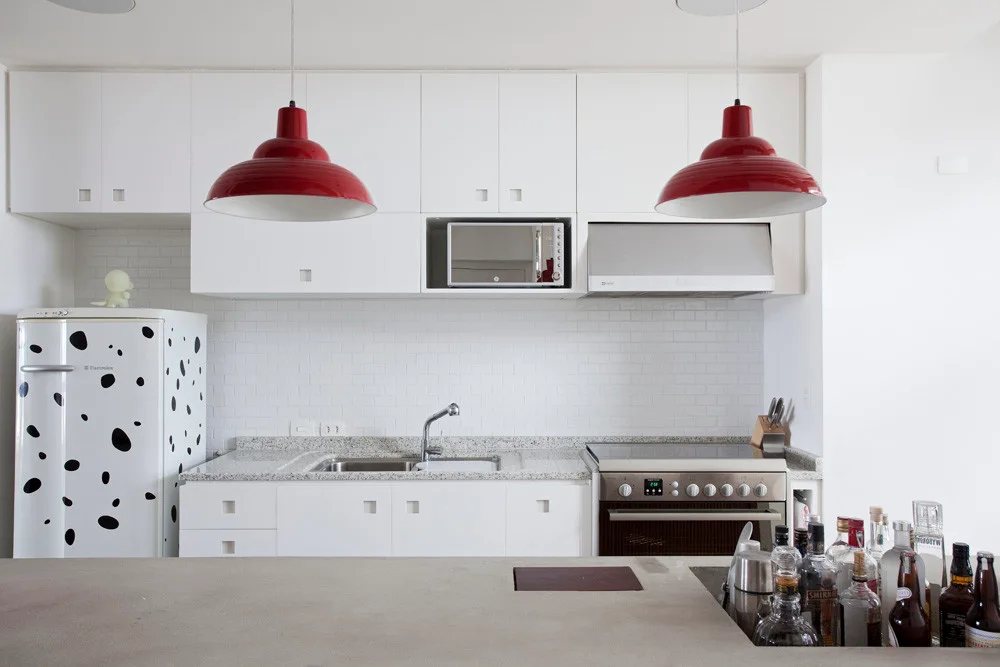 A modern white kitchen features flat-front cabinetry, white subway tiles, a spotted refrigerator, and two bold red dome pendant lights over the counter.