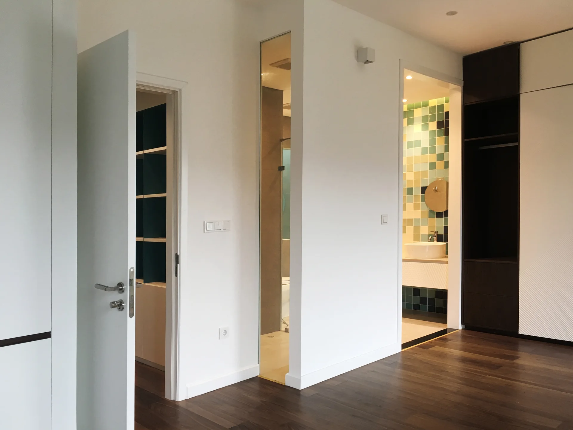 An open doorway in a white bedroom reveals a modern bathroom featuring a colorful green and yellow tiled wall, a vessel sink, and a round mirror.