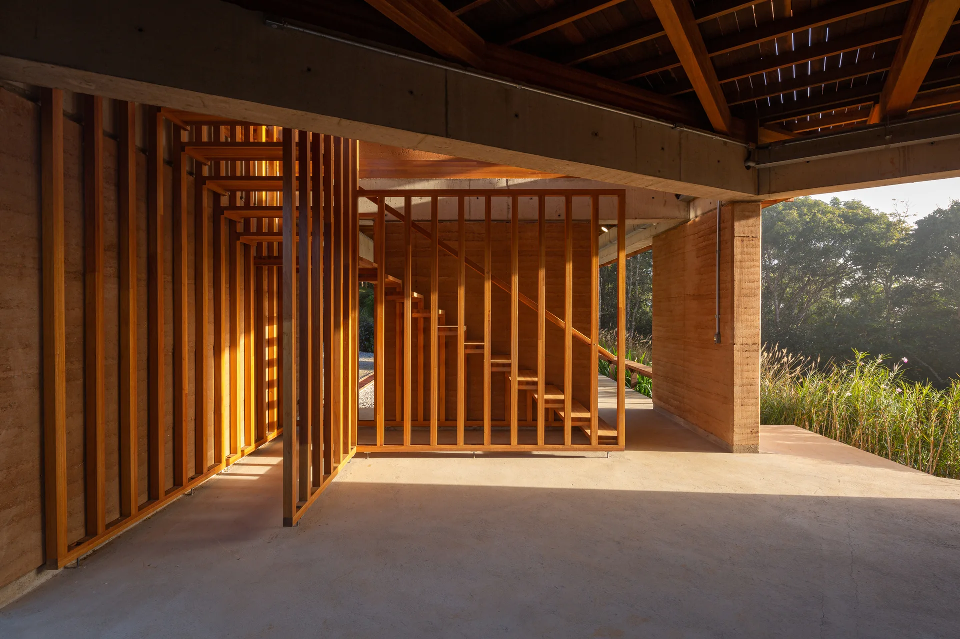 A wooden staircase with a vertical slat partition is set against textured earth walls and an open transition to the grassy outdoors.