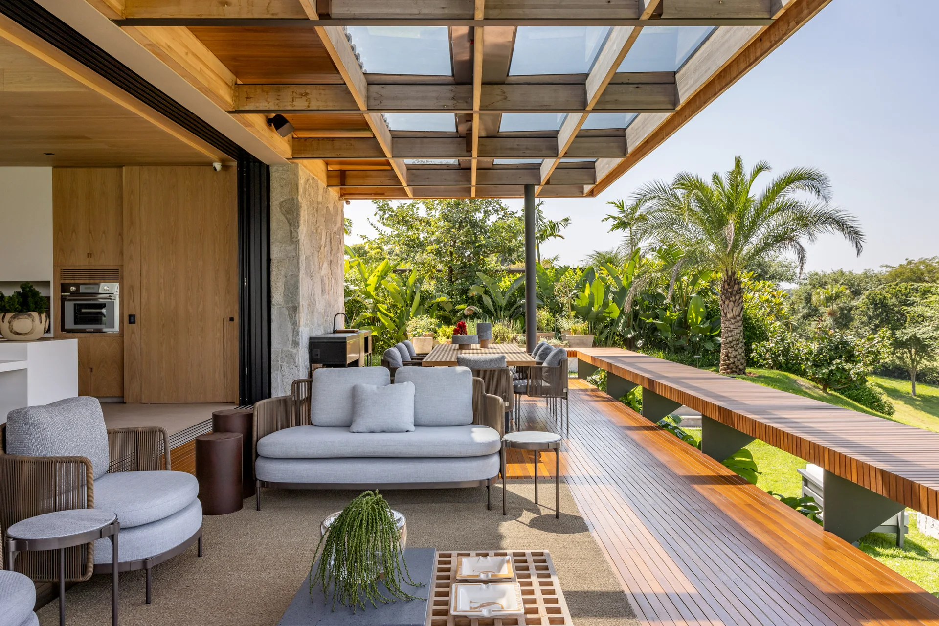 A covered outdoor patio featuring a timber pergola, gray upholstered lounge furniture, and a wood dining table overlooking a lush garden.