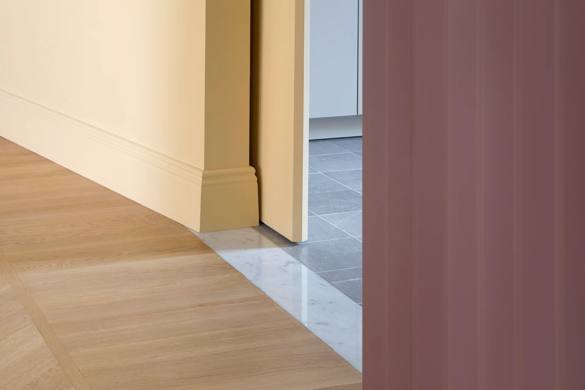 A close-up view of a doorway transition showing light wood flooring, a white marble threshold, grey tile, and contrasting yellow and mauve walls.