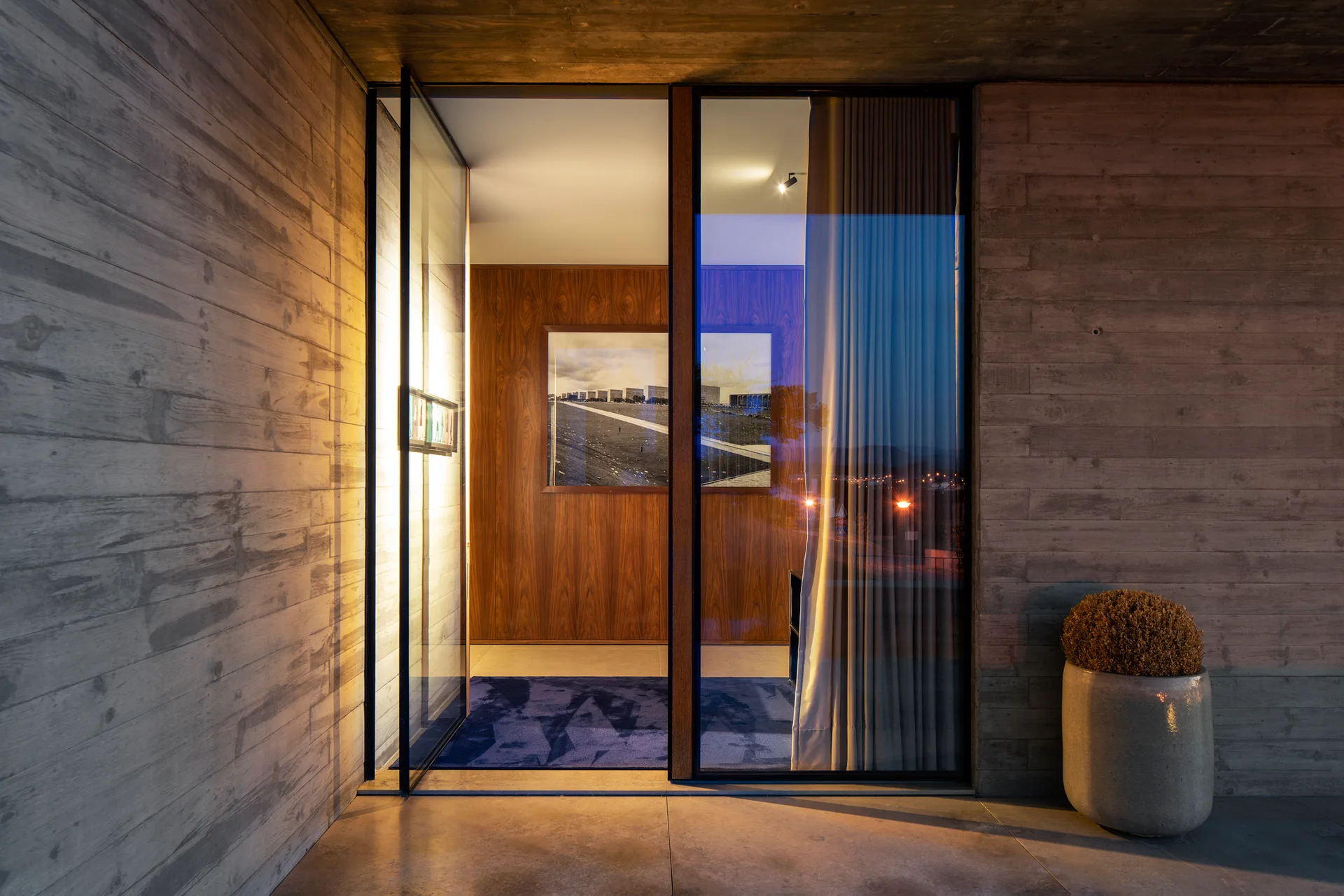 A modern entryway with board-formed concrete walls and a glass pivot door opening into a foyer with warm wood paneling and a blue rug.