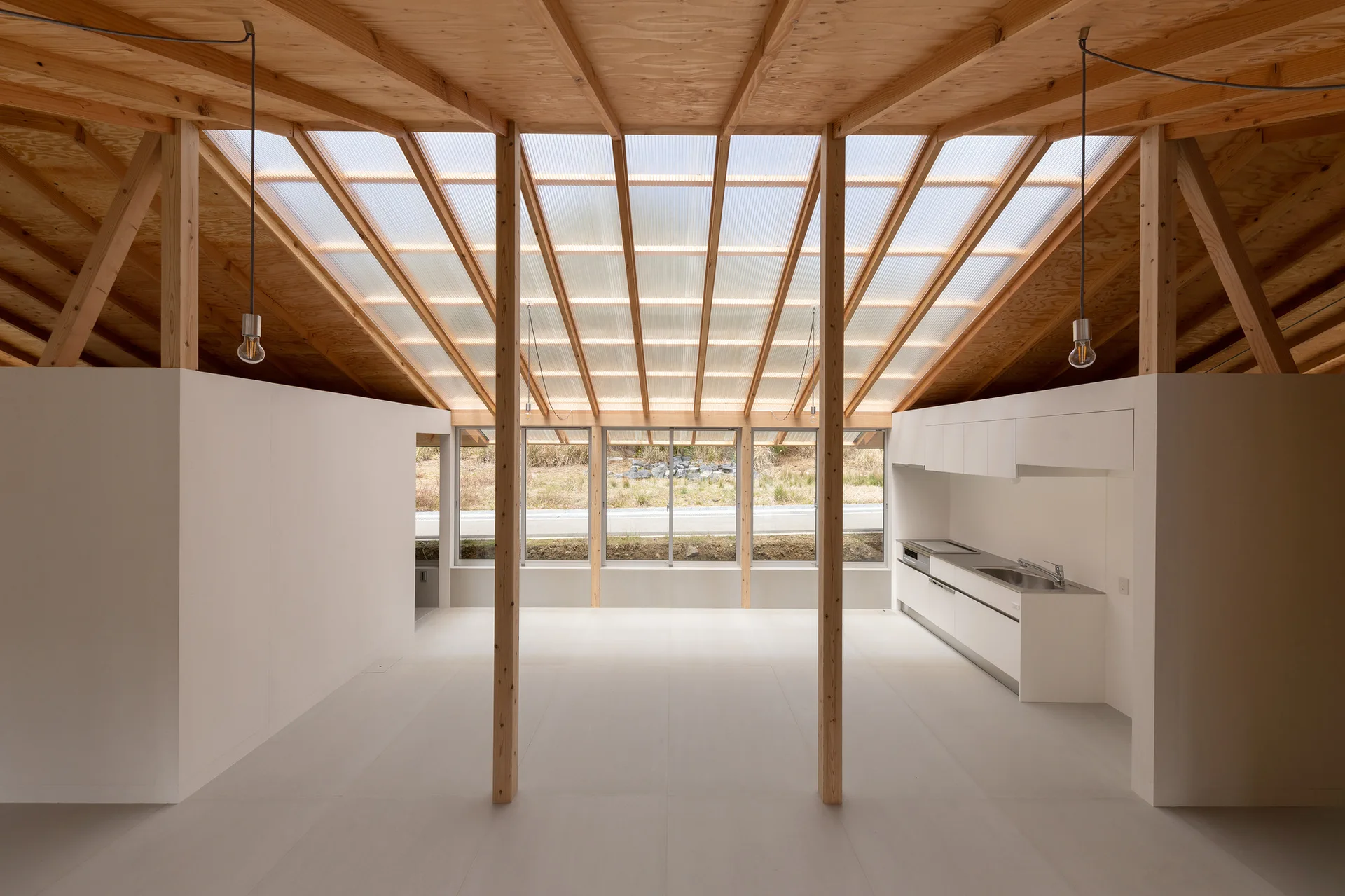 A minimalist open-plan kitchen with white cabinetry, exposed wooden support pillars, and a sloped ceiling featuring translucent skylight panels.