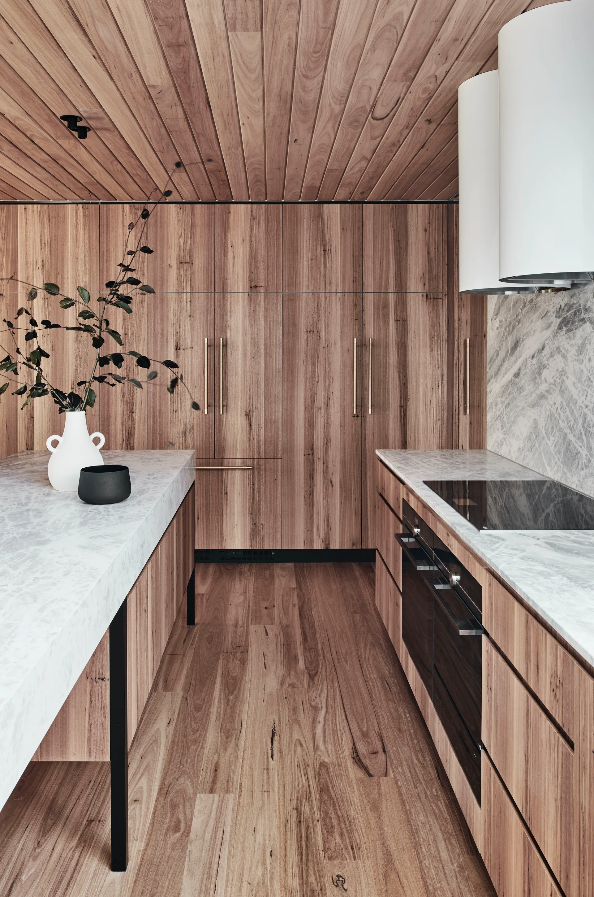 A modern galley kitchen features extensive wood-grained cabinetry, matching wood plank ceilings, and grey-veined white marble countertops.