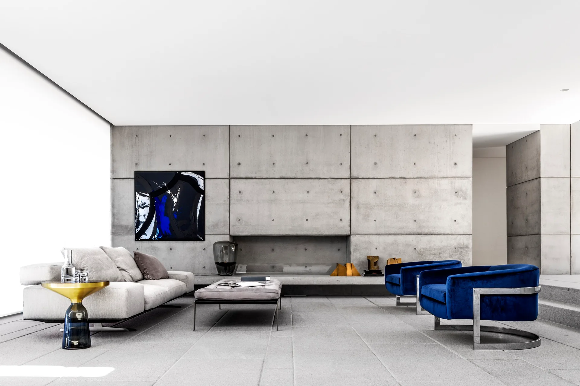 A modern living room features a concrete panel wall, a light gray sofa, and two deep blue velvet armchairs on a light gray stone tile floor.