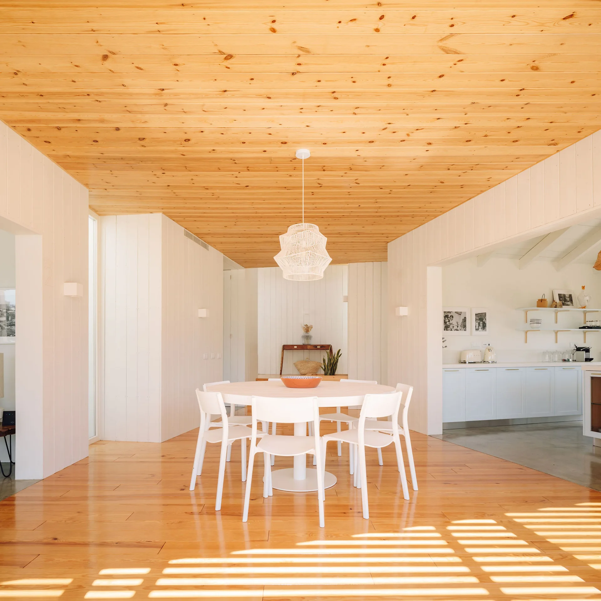 A bright dining room featuring a white circular table, matching chairs, vertical wood-paneled walls, and a natural pine ceiling.