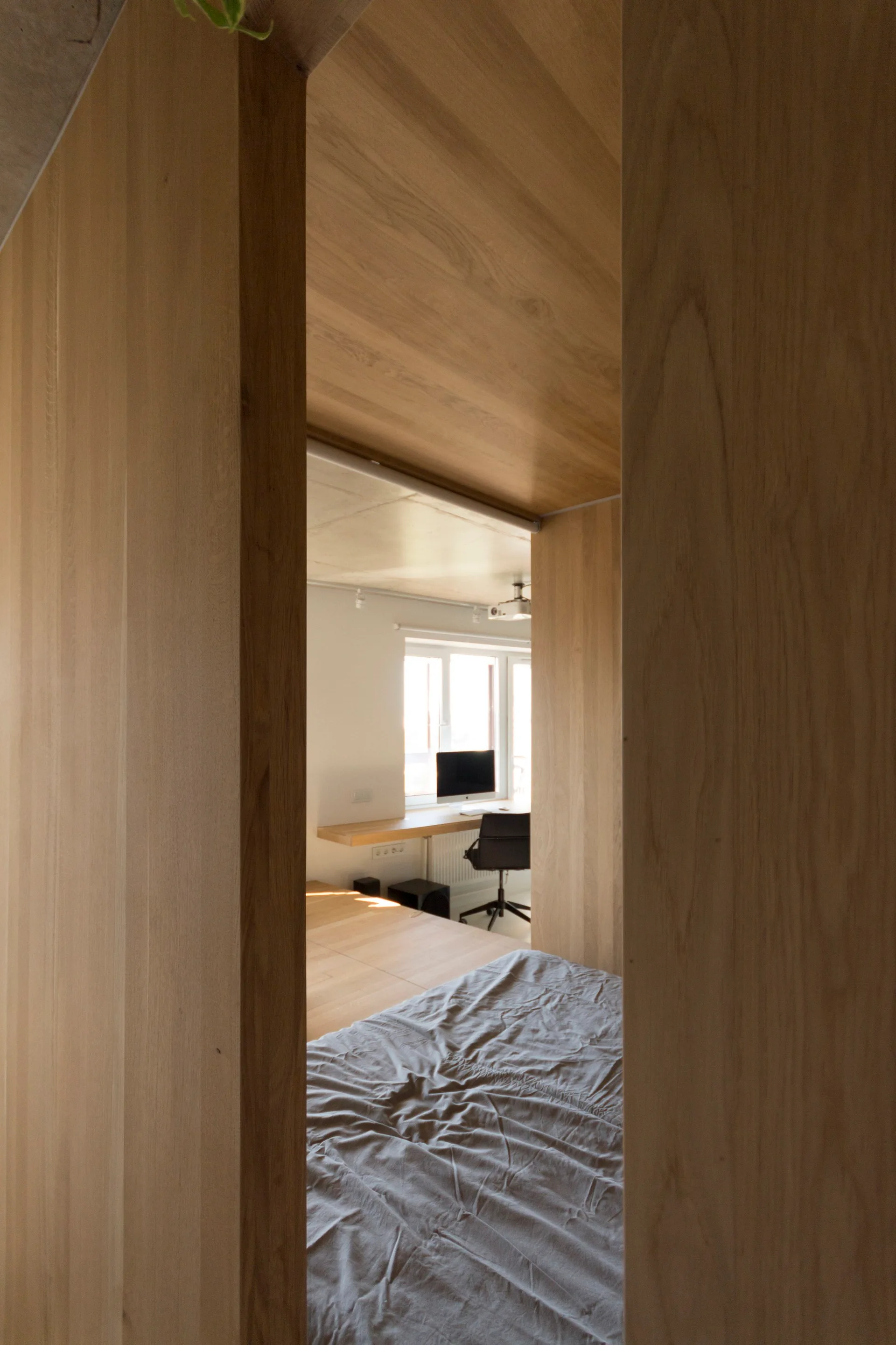 A view through a natural wood partition looking from a bed with grey sheets toward a minimalist home office nook with a floating desk and window.