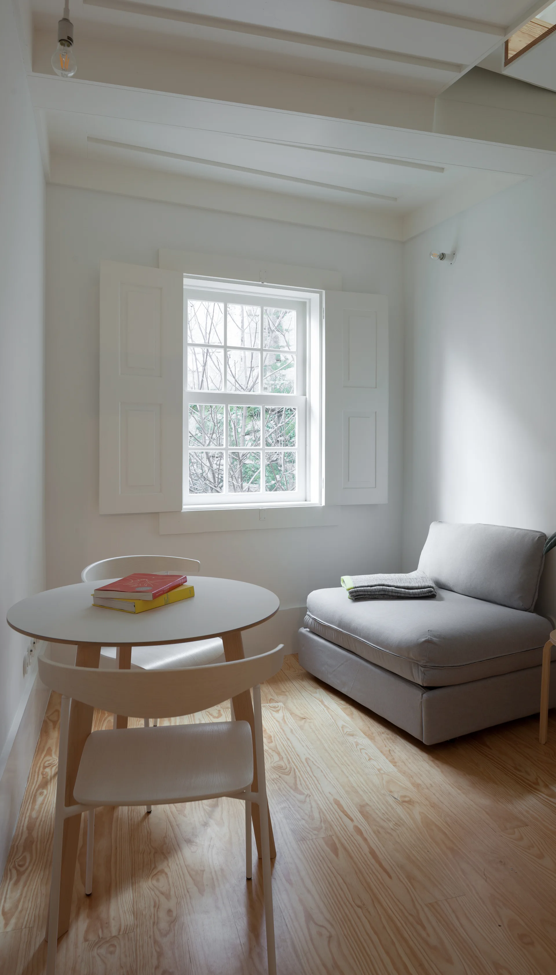 A minimalist room with a round wooden table, two chairs, a grey armless lounge chair, and white interior shutters on the window.