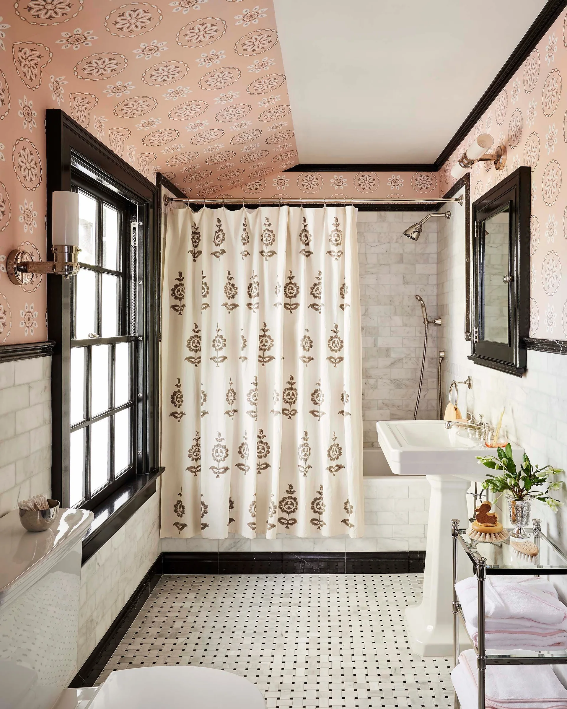 A traditional bathroom featuring pink floral wallpaper, high-contrast black trim, a white pedestal sink, and marble wall tiles.