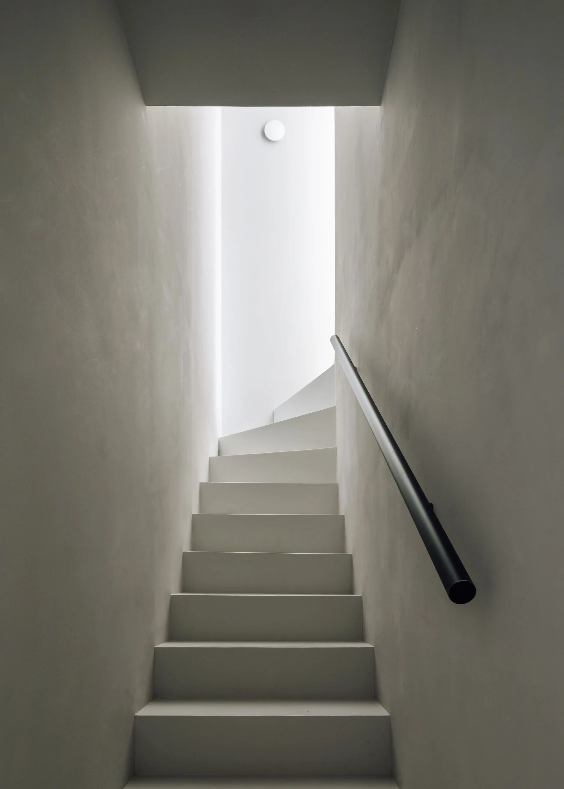 A minimalist white staircase ascends between smooth light gray plaster walls toward a bright ceiling light with a simple black handrail on the right.