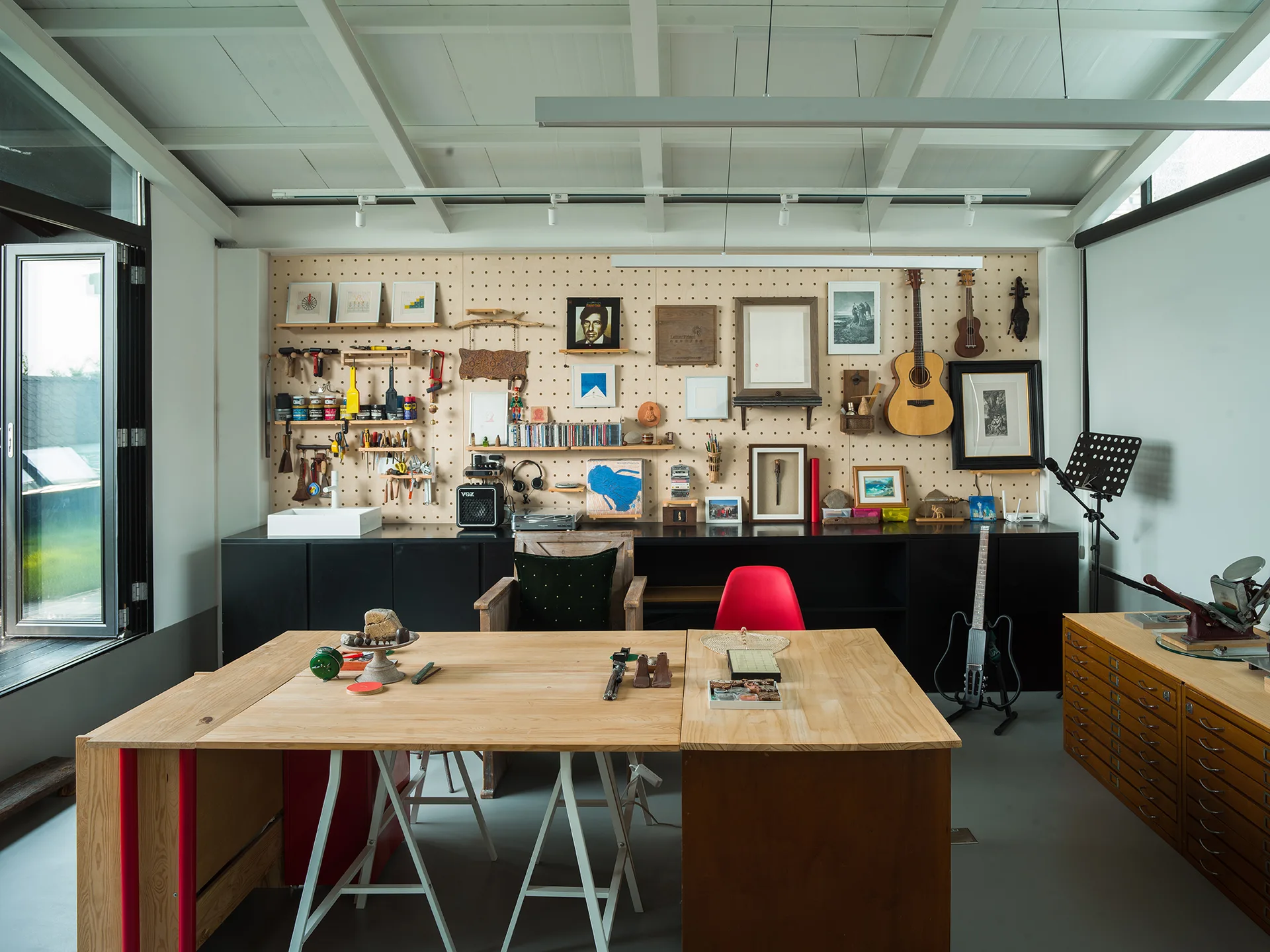 A bright creative studio featuring a large wooden work table, a full-wall pegboard storage display, and sleek black cabinets.