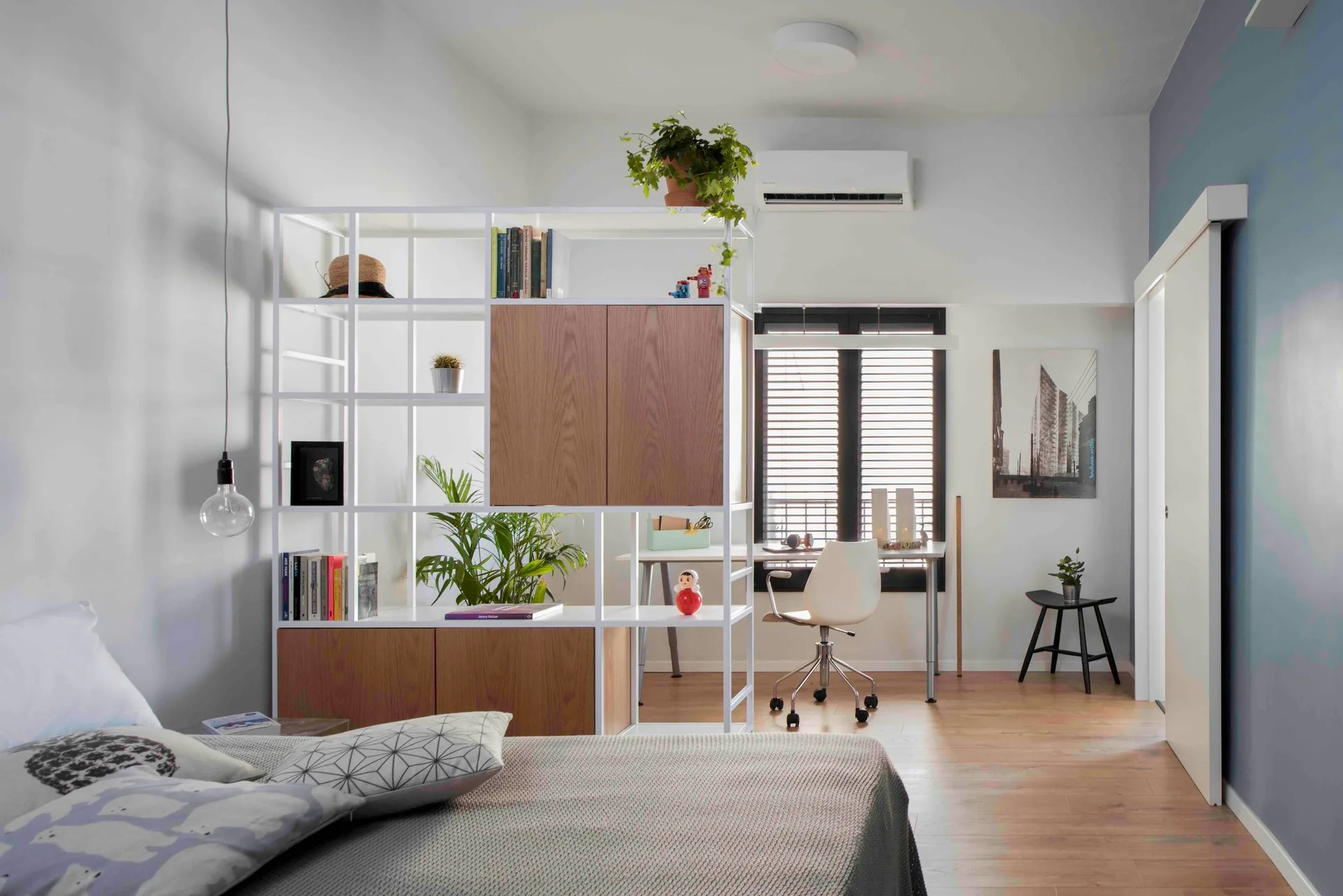 A white open shelving unit serves as a room divider between a bed with patterned grey pillows and a home office desk with a white chair.