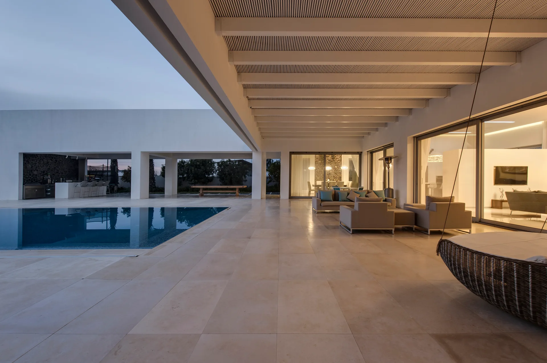 A modern covered patio features a beige modular sofa and hanging chair next to a rectangular swimming pool under a white slatted ceiling.