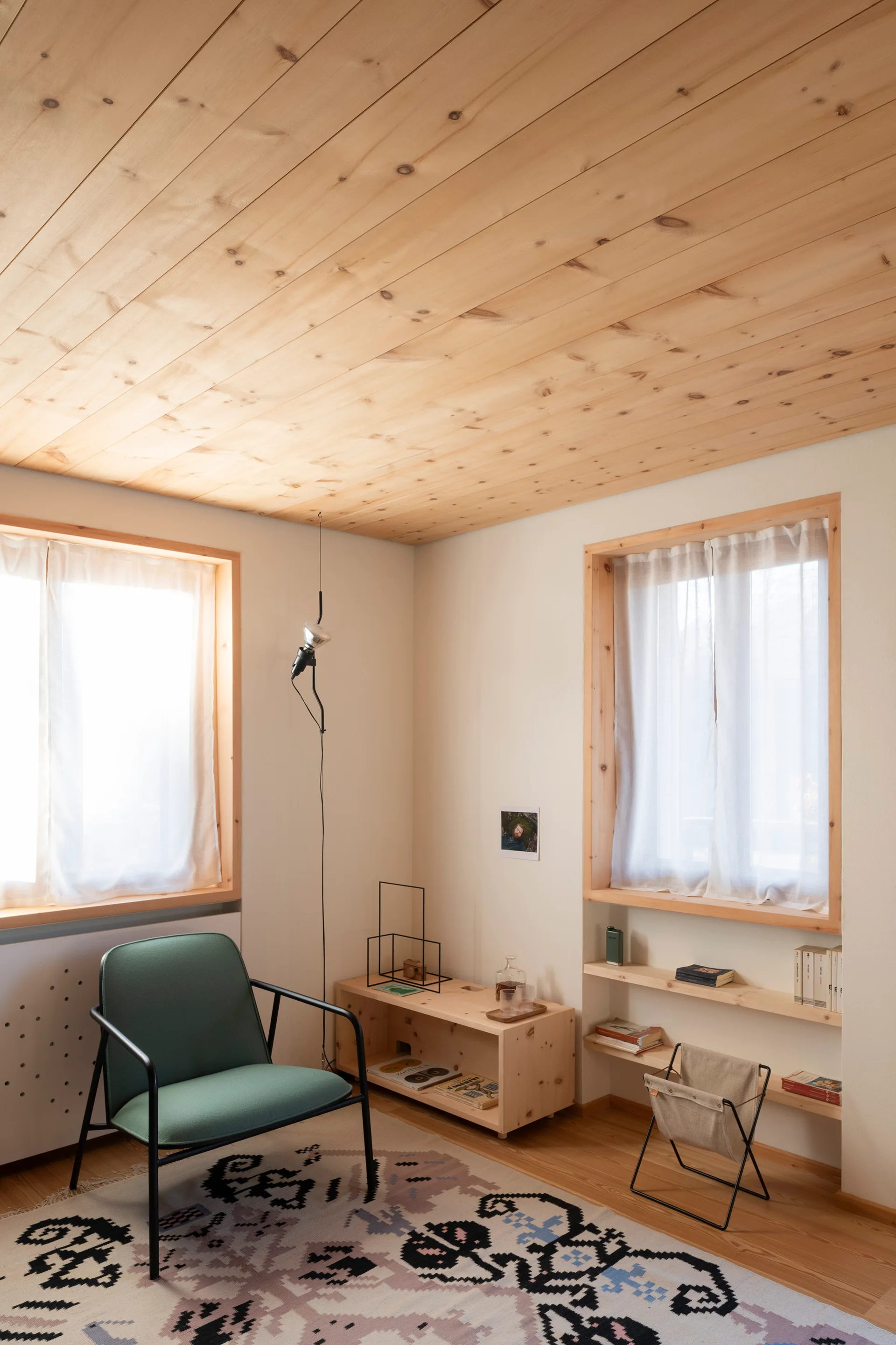 A minimalist living room corner features a green armchair, a low wood storage bench, and sheered windows under a planked pine wood ceiling.
