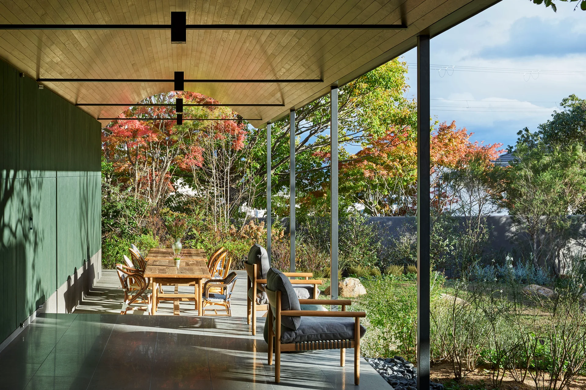 A covered outdoor patio features a curved wood plank ceiling, a wooden dining table with chairs, and grey lounge chairs overlooking a garden.
