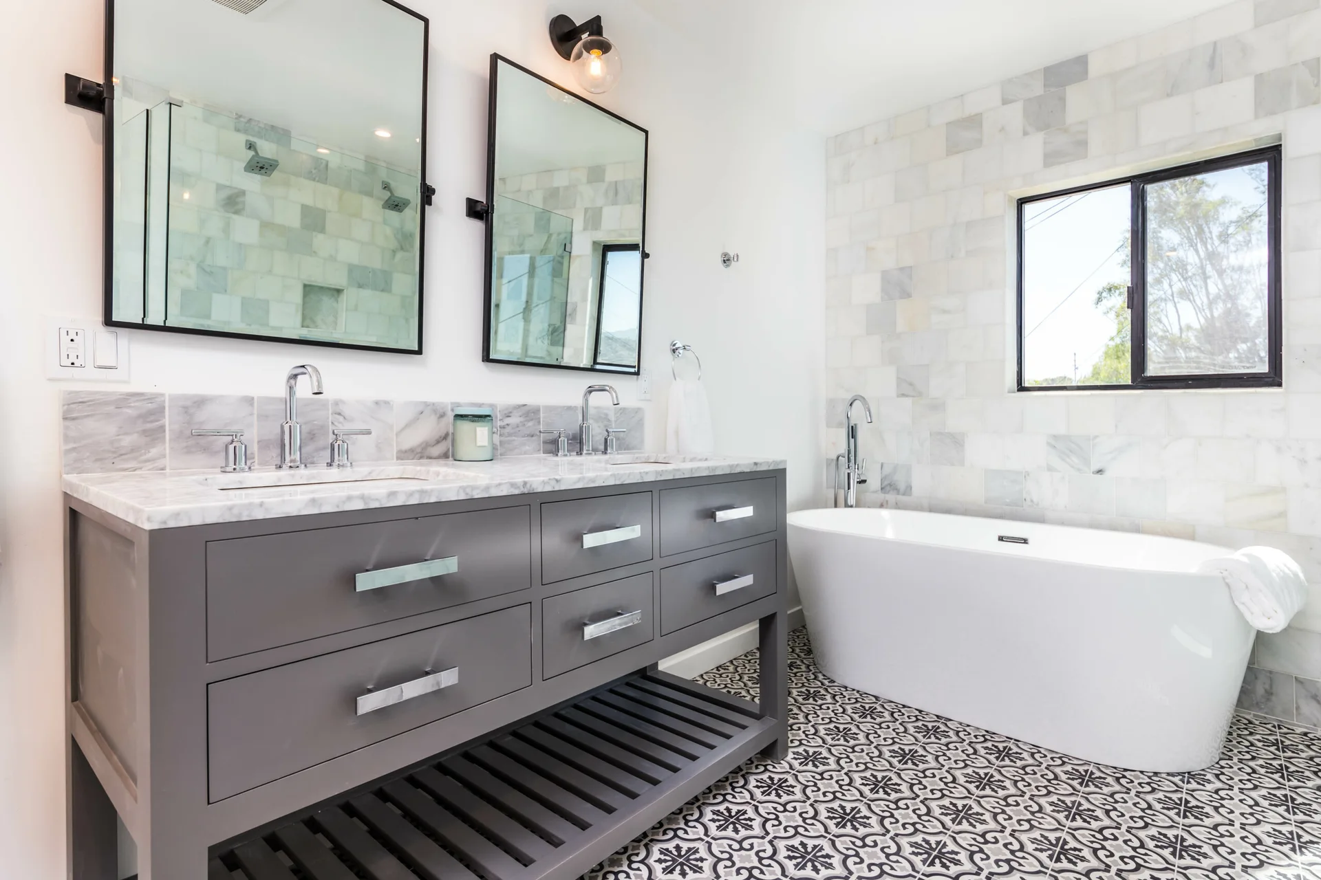 A modern bathroom featuring a grey double vanity, two black-framed mirrors, a freestanding white bathtub, and patterned black and white floor tiles.