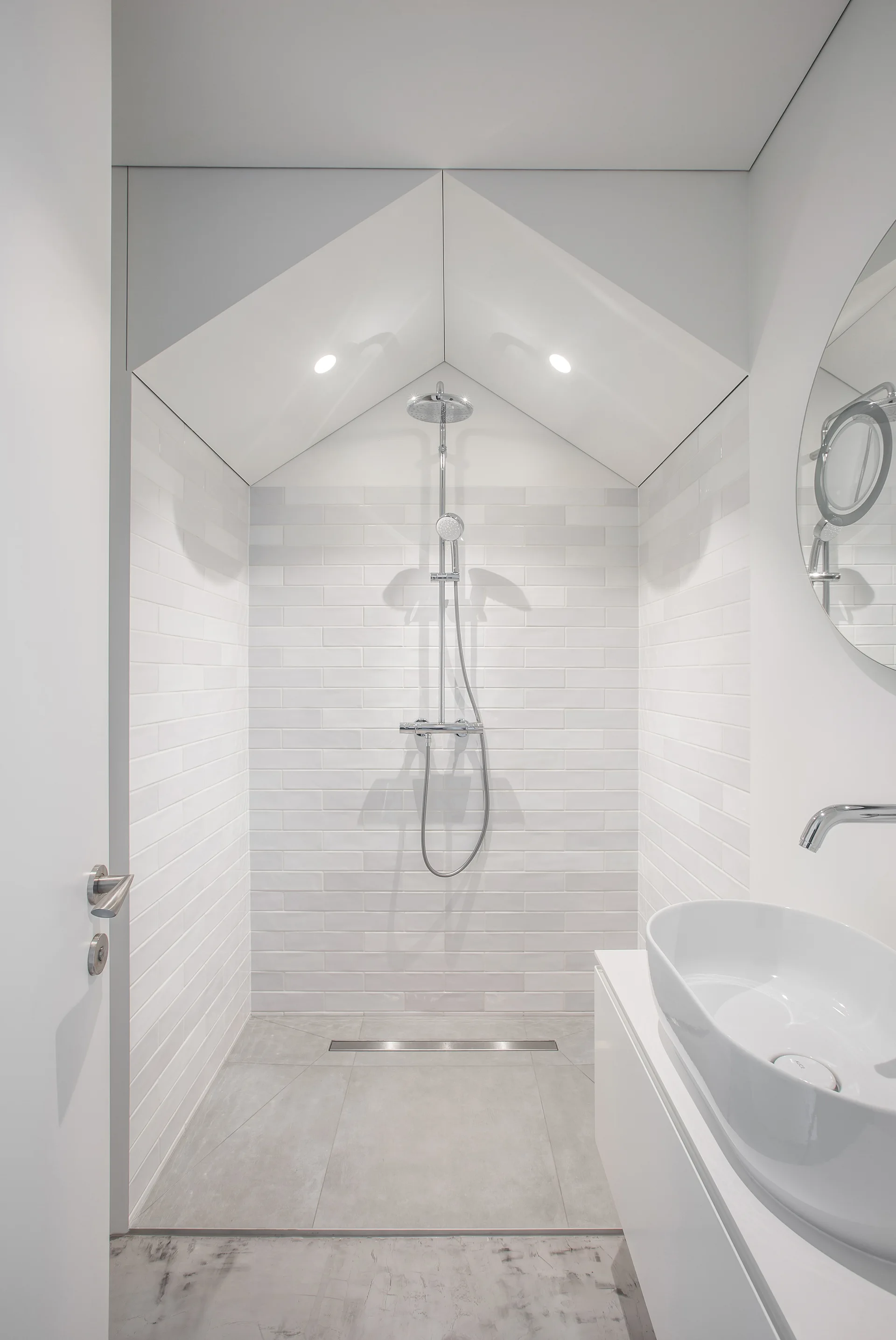 A minimalist white bathroom with a walk-in shower featuring a pitched gable ceiling, glossy white subway tile walls, and a white vessel sink.