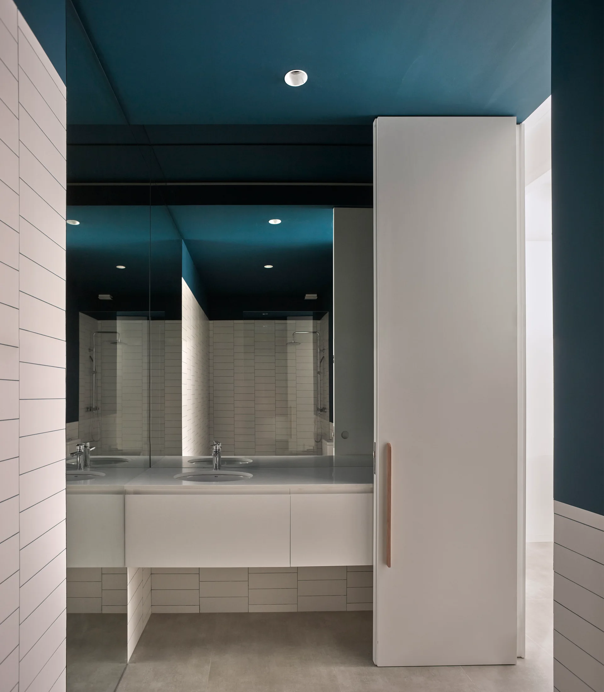 A minimalist bathroom features a white wall-mounted vanity, white tiled walls, a large mirror, and a deep blue painted ceiling.