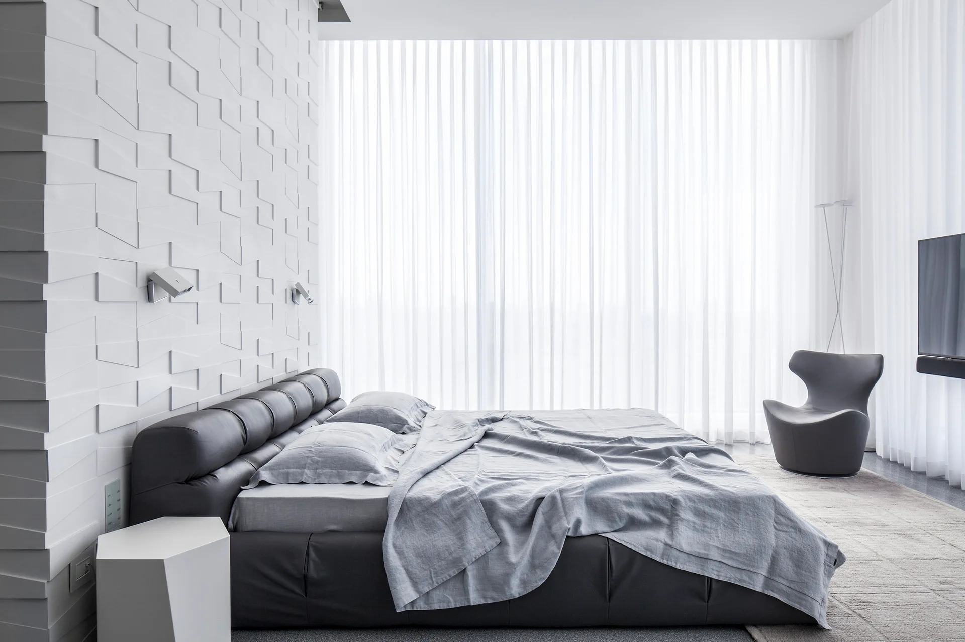 A modern bedroom features a black leather bed set against a textured white geometric wall with sheer curtains in the background.