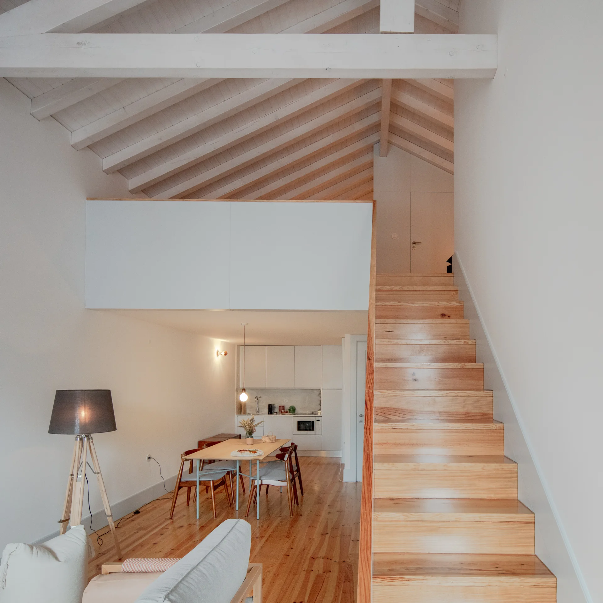 A wooden staircase leads to a loft mezzanine in an open-plan living area with exposed ceiling beams, a dining set, and a compact white kitchen.