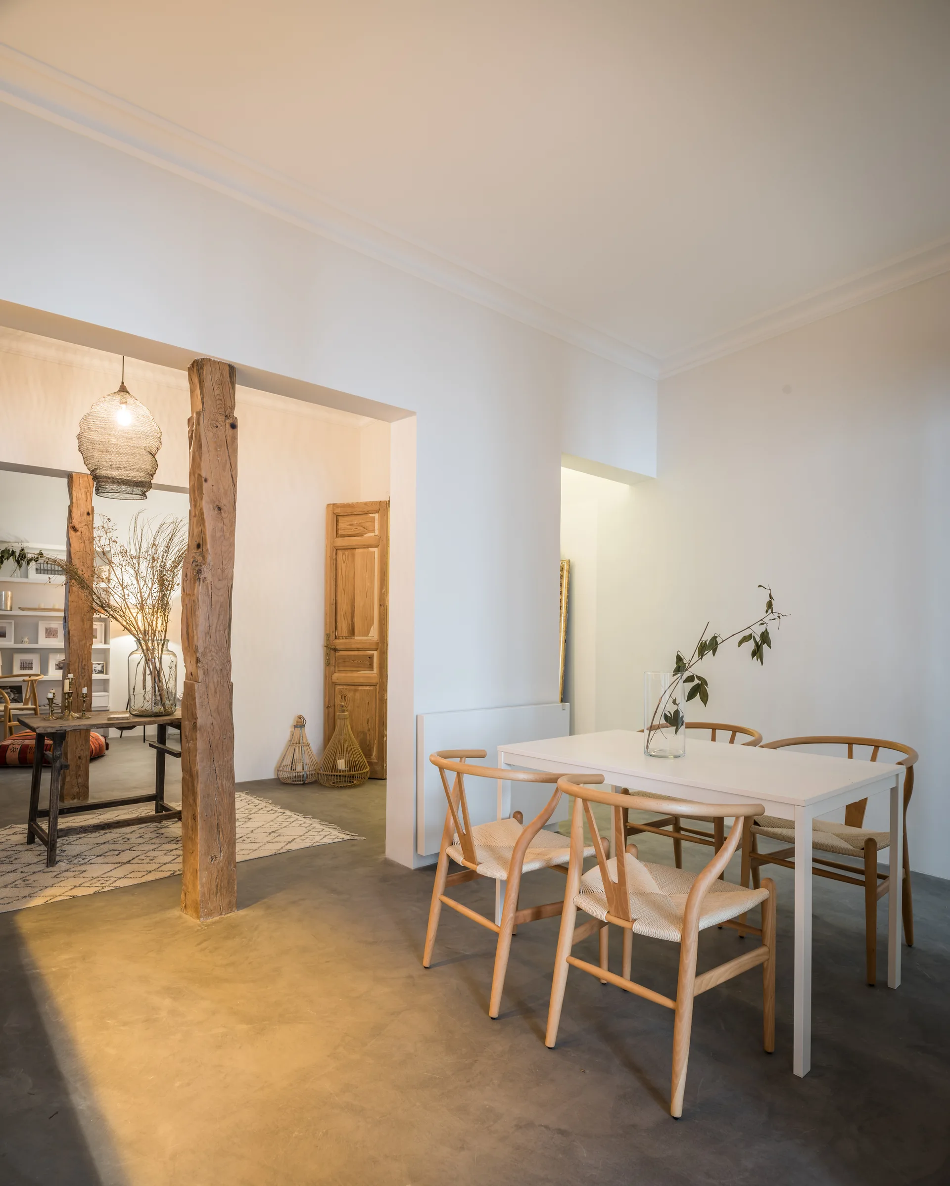 A minimalist dining room features a white table with light wood wishbone chairs set near a rustic wood support column and grey concrete floors.