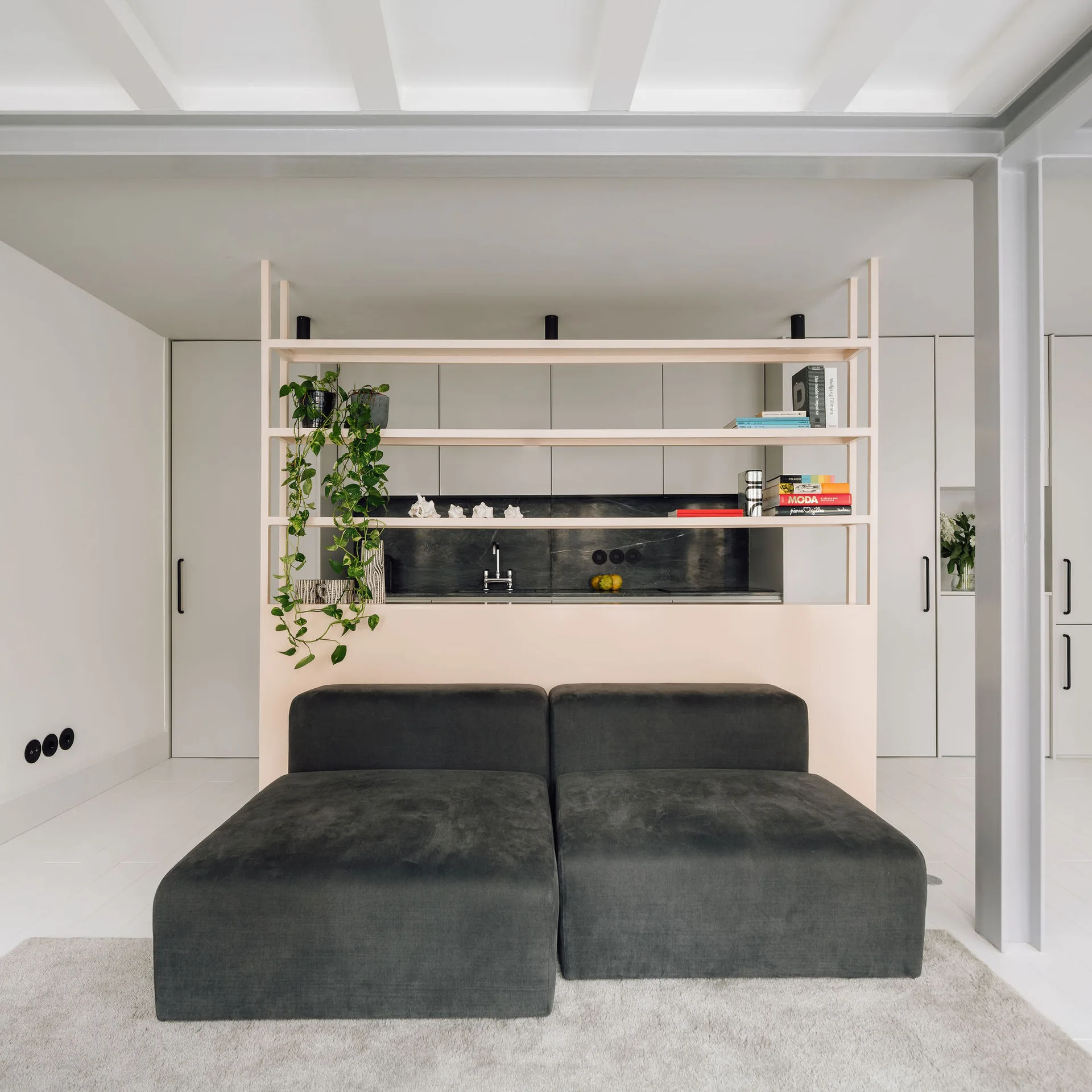 A low charcoal modular sofa sits in front of a pale wood room divider shelf that separates the living area from a kitchen with dark countertops.
