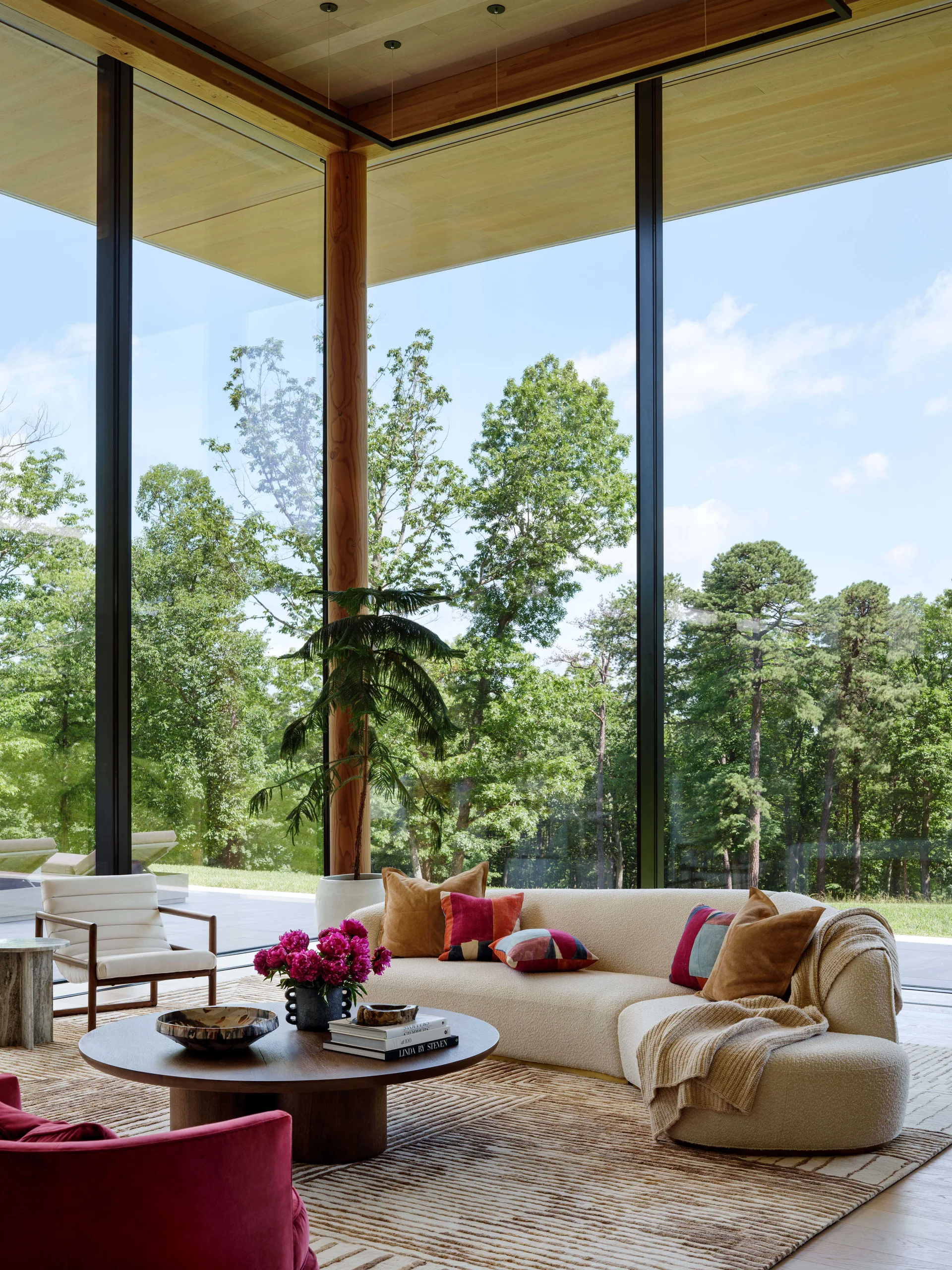 A contemporary living room featuring a curved cream sofa, wooden coffee table, and textured rug positioned before floor-to-ceiling glass walls.