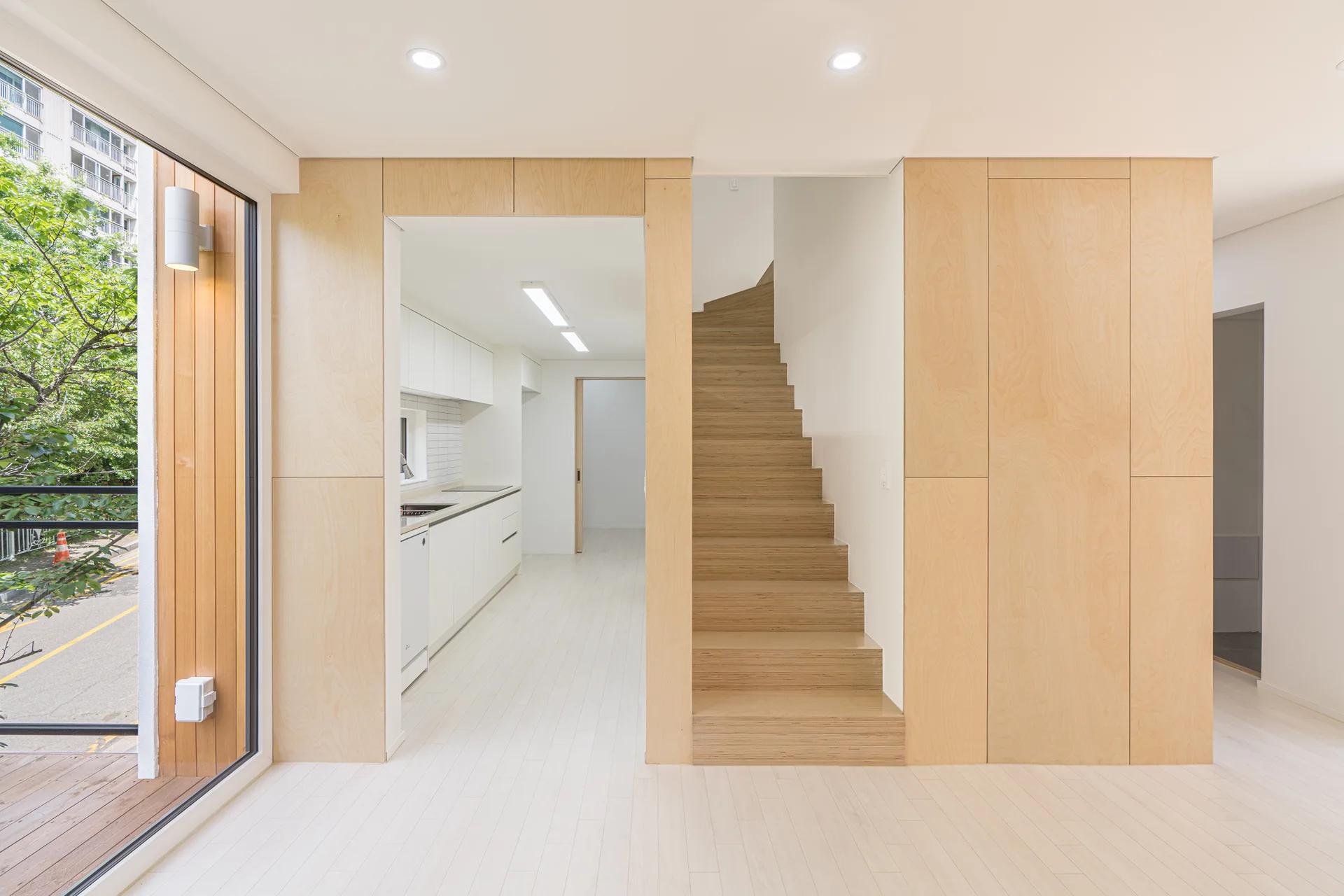 A minimalist interior featuring a central light-wood staircase framed by matching plywood wall panels, with a white kitchen visible in the background.