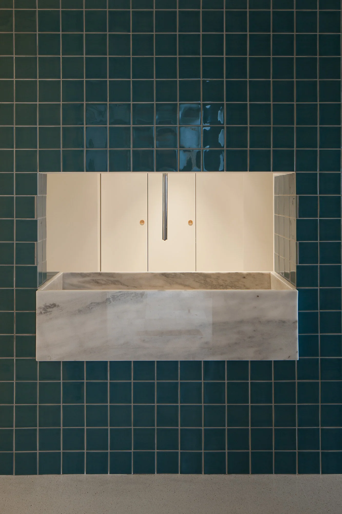 A modern wall-mounted marble sink is set against a glossy teal tiled wall featuring a recessed niche with white cabinets and a chrome faucet.