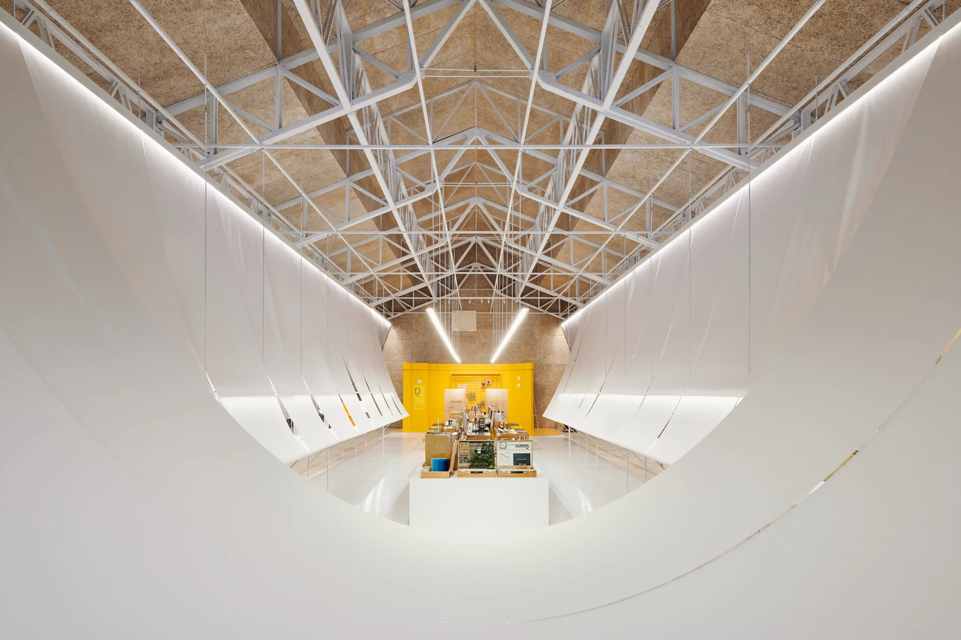 A large industrial hall featuring exposed metal ceiling trusses, a curved white canopy installation, a glossy floor, and a bright yellow back wall.