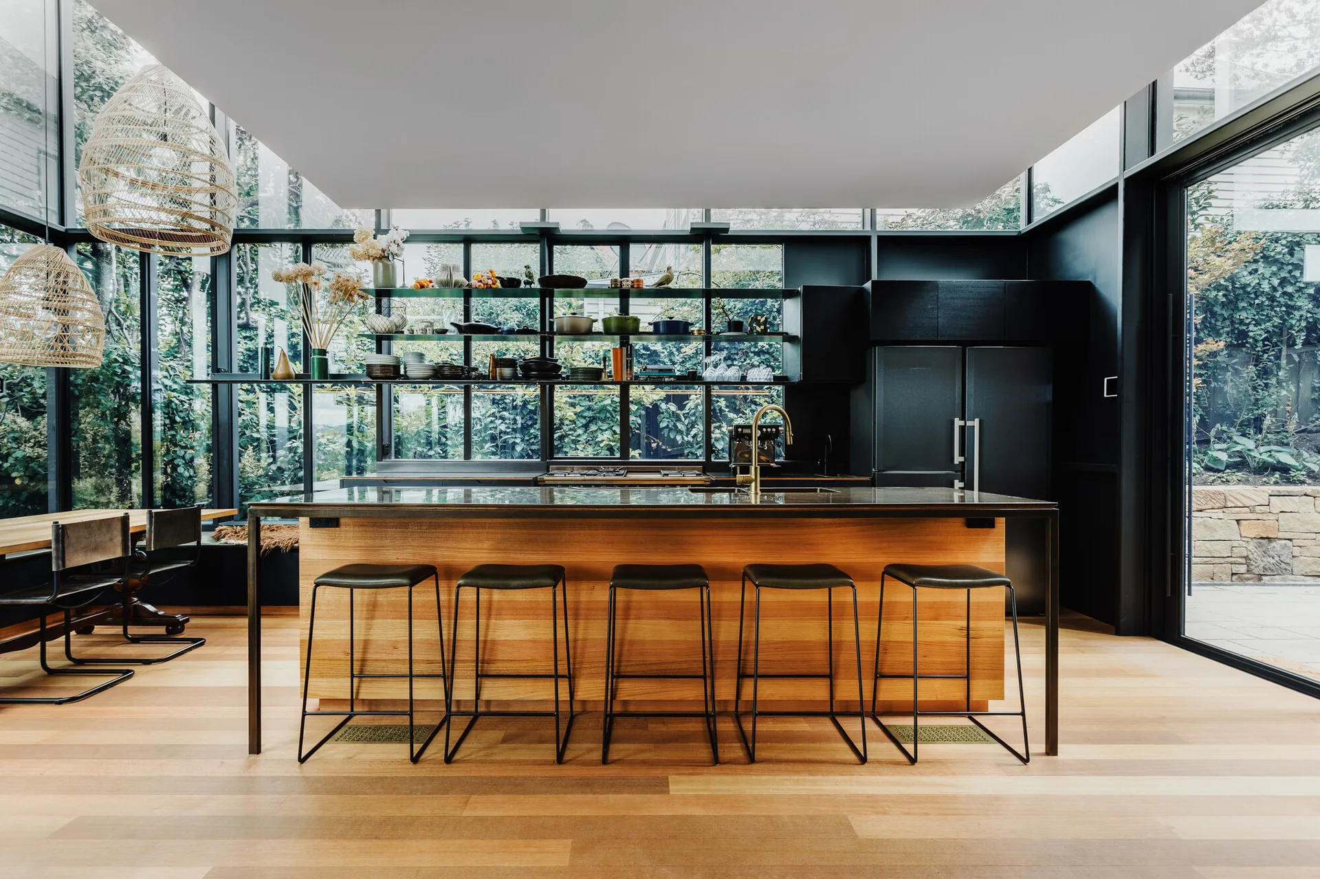 A modern kitchen with a large wood island, black bar stools, and open shelves displaying kitchenware against a wall of black-framed glass windows.