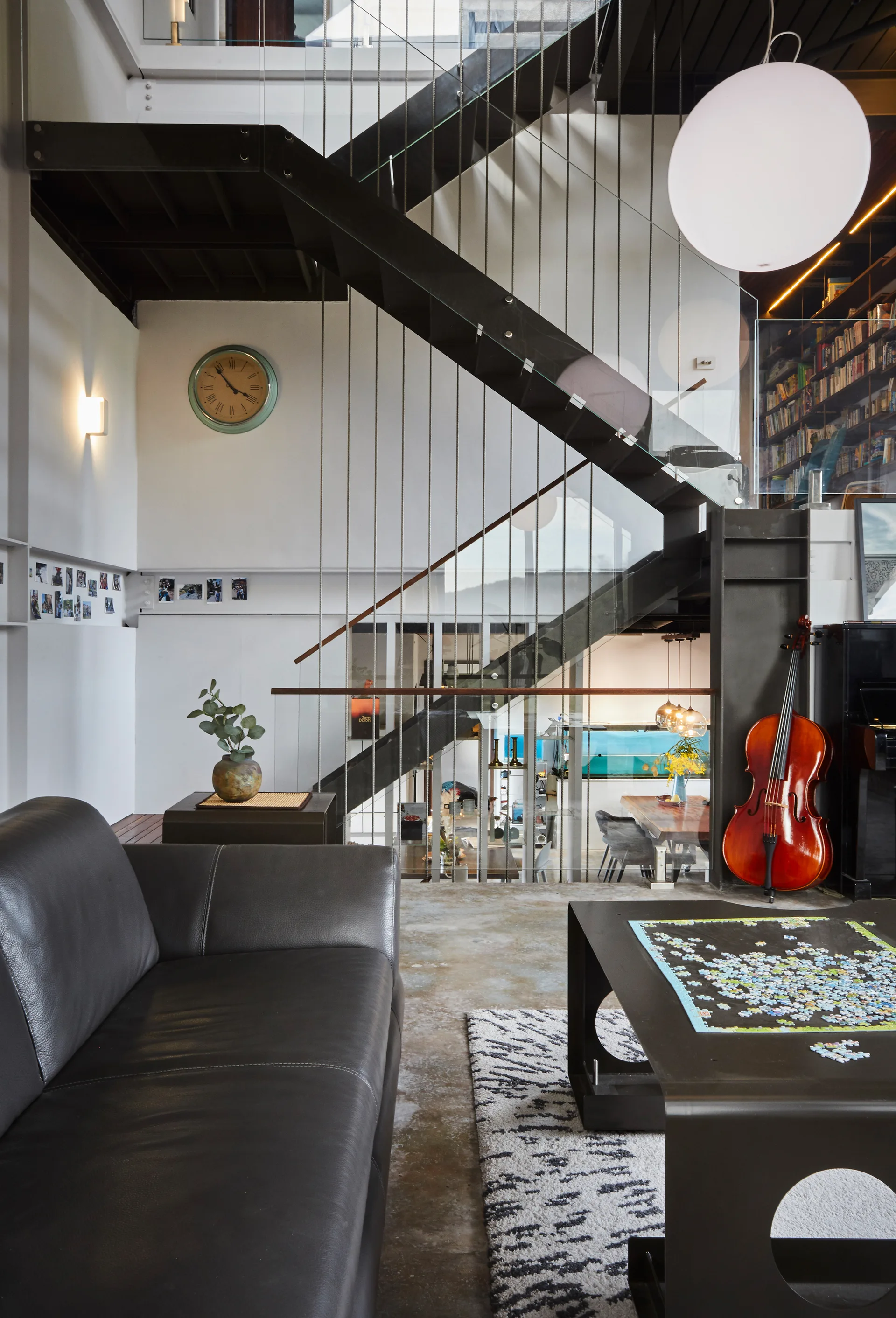An industrial loft living space with a black metal staircase, glass railings, concrete floors, a dark leather sofa, and a wooden cello in the background.