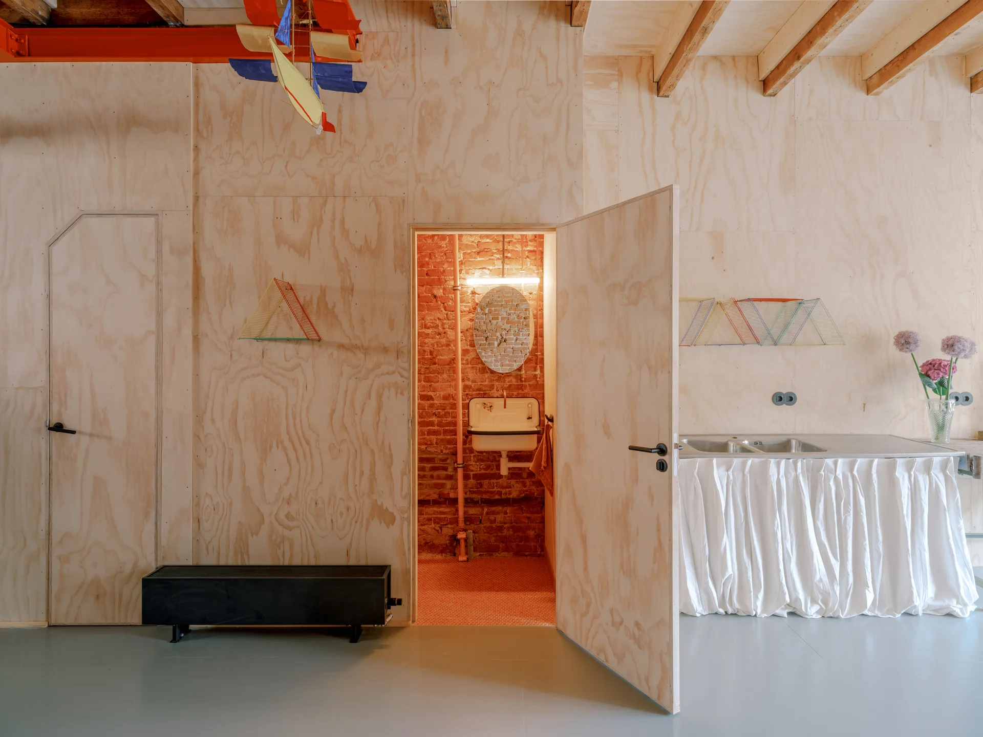 An open plywood door reveals a bathroom with exposed brick walls, a vintage sink, and an oval mirror next to a kitchen sink with a fabric skirt.