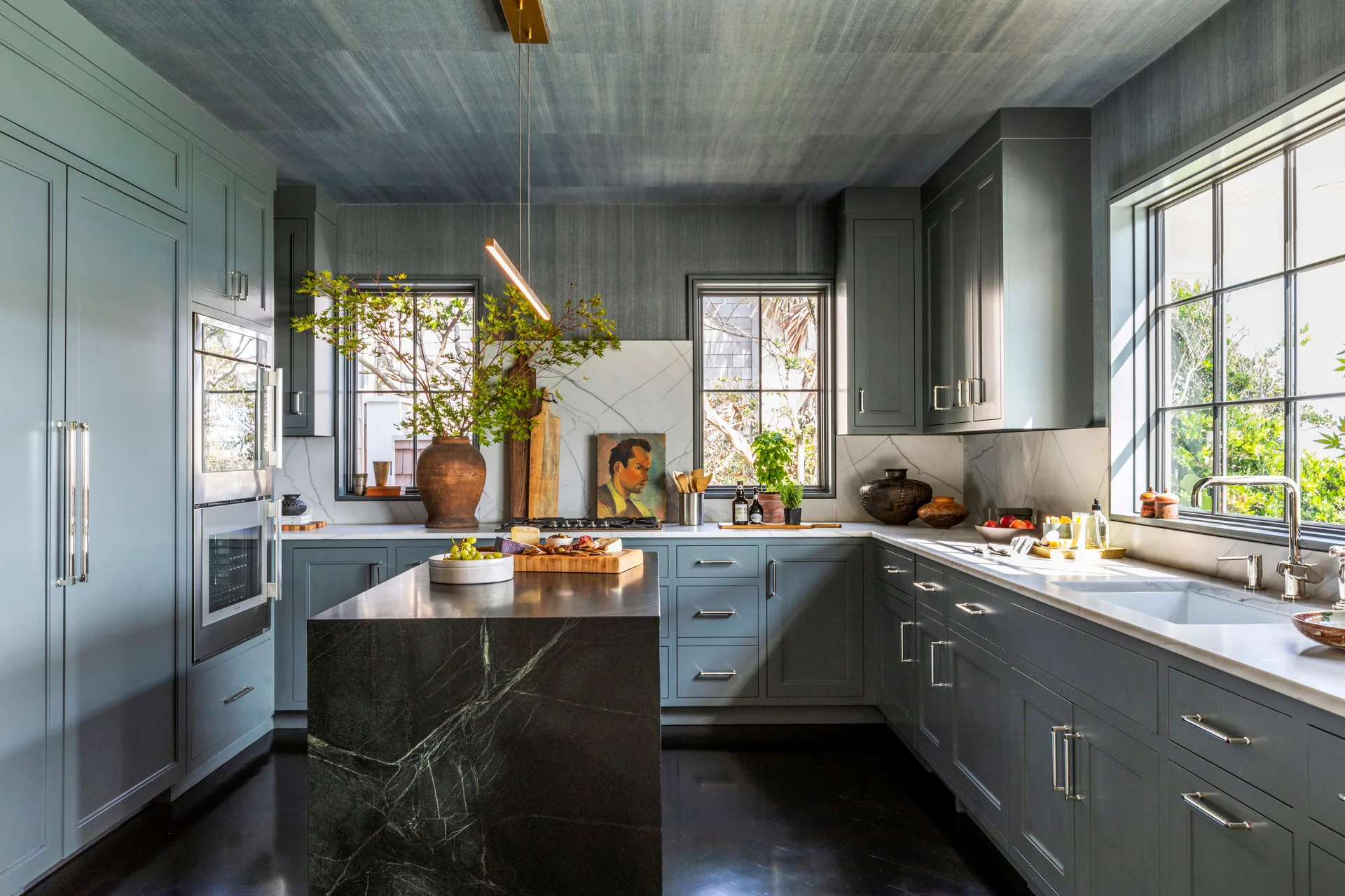 Gray shaker cabinets surround a dark marble kitchen island under a textured wallpapered ceiling with two multi-pane windows.